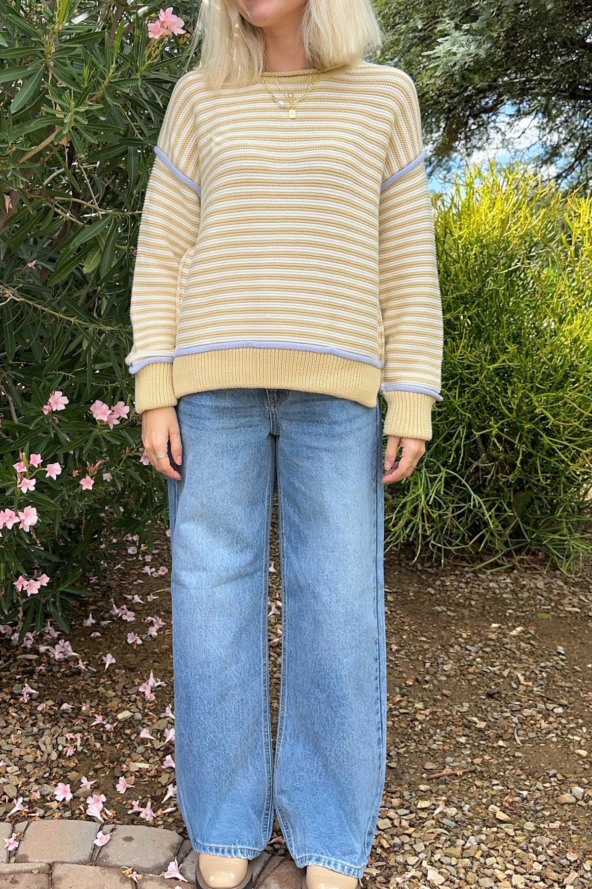 A person standing outdoors in front of green bushes and flowers, wearing a cream and tan striped knit sweater with light blue wide-leg jeans.