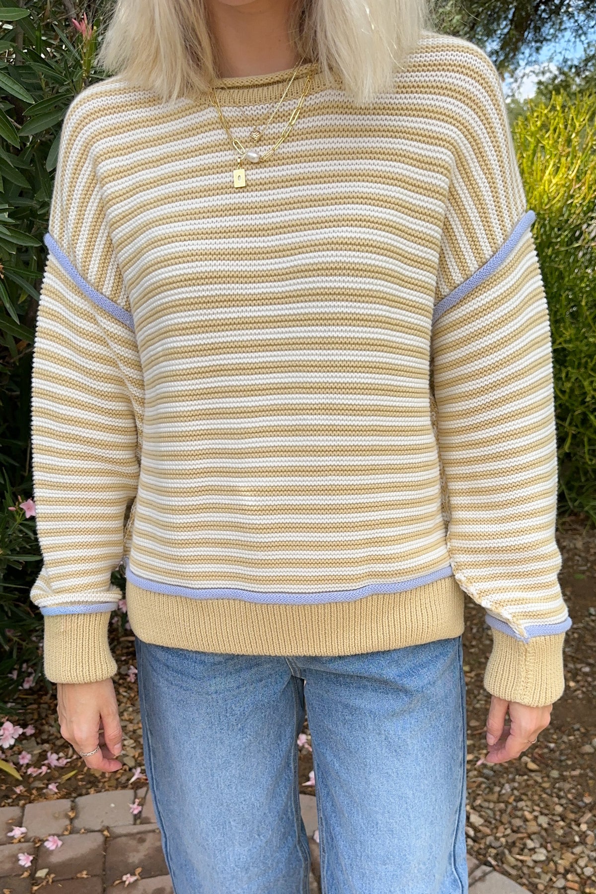 A person wearing a cream and tan striped knit sweater with light purple contrast trim on the shoulders and cuffs, paired with light blue jeans, photographed outdoors in front of greenery.