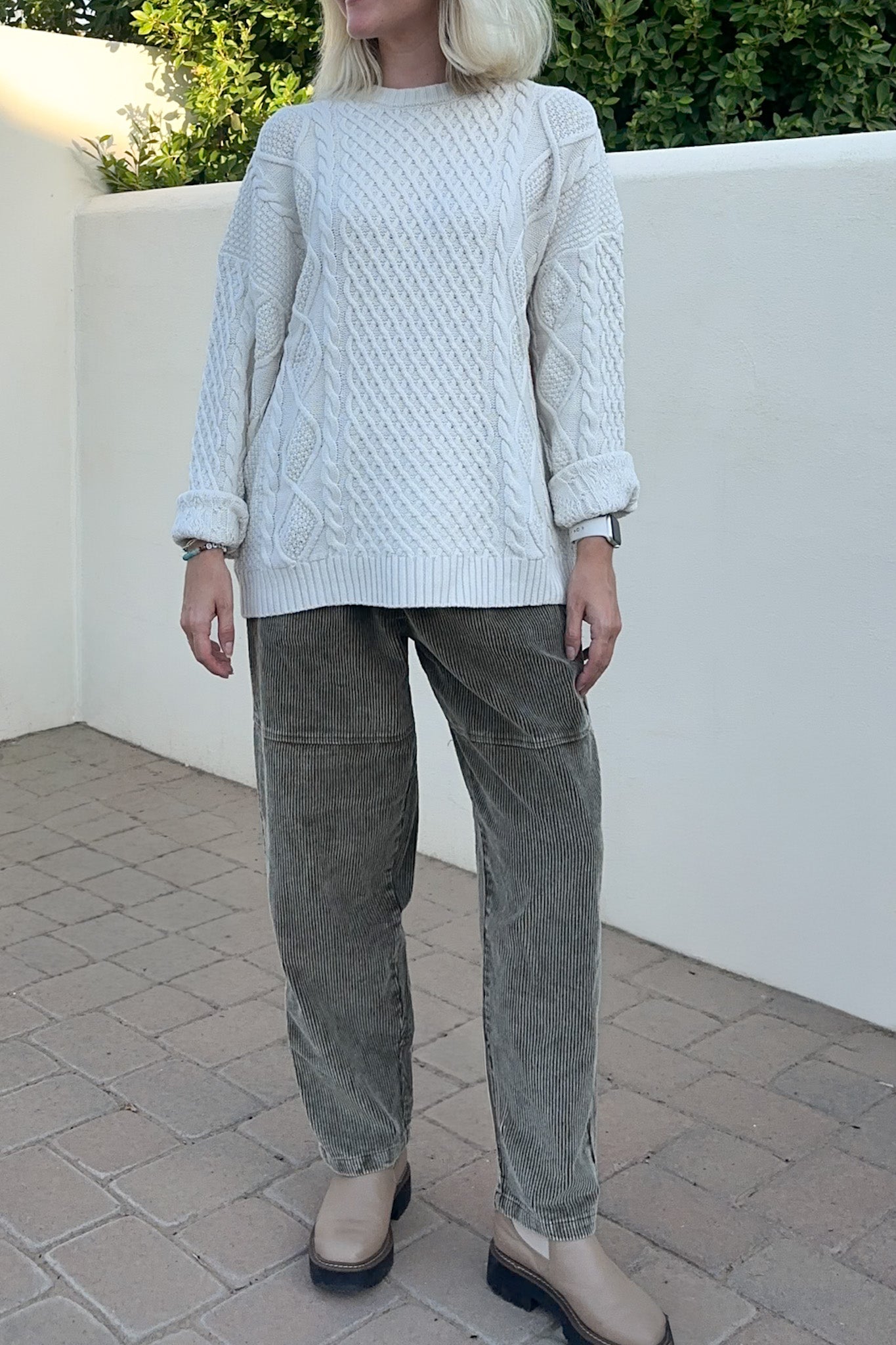 A woman wearing a white cable-knit sweater and olive green corduroy pants, standing outdoors against a white wall.
