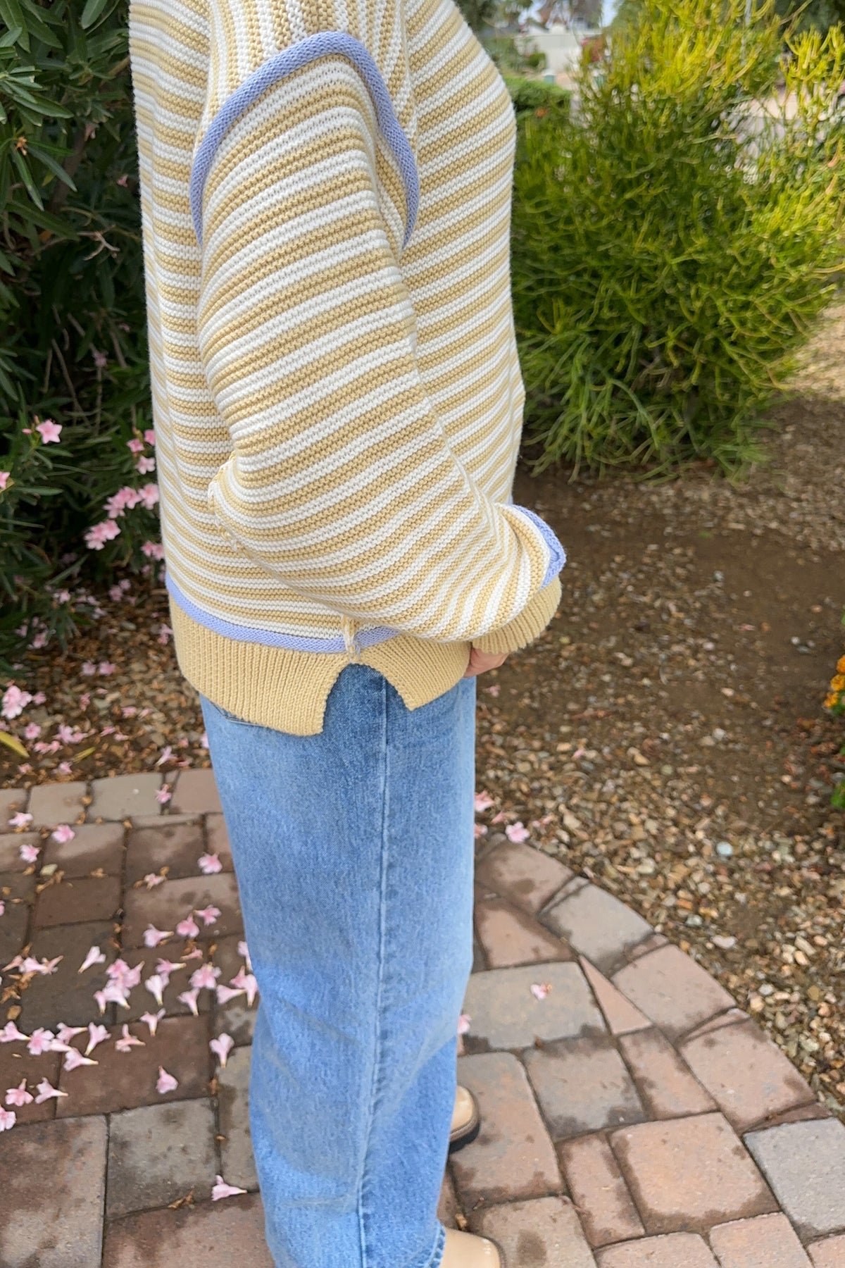 A person outdoors wearing a cream and yellow striped knit sweater with light blue trim, paired with light blue jeans, standing on a brick path with garden plants in the background.