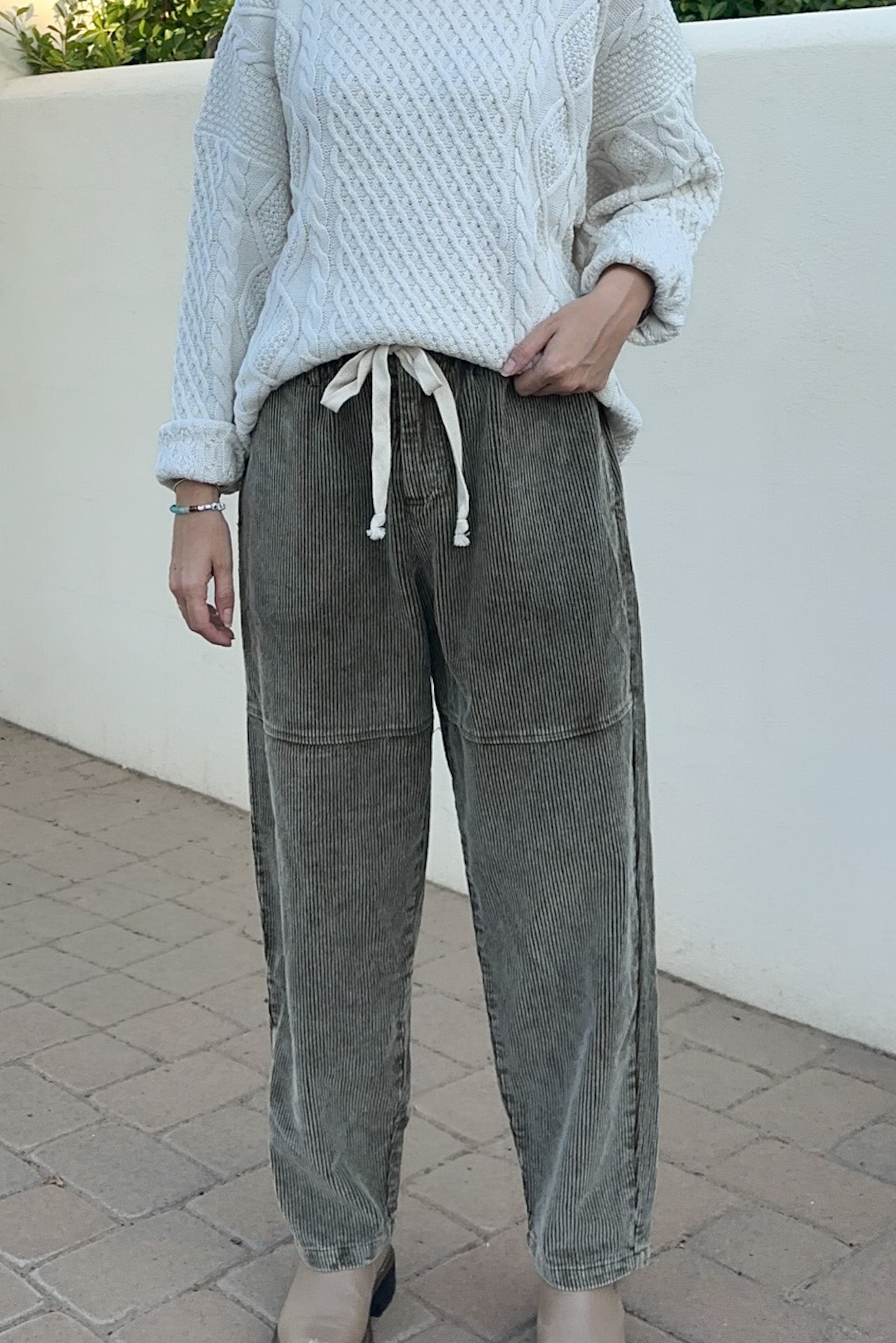 A person wearing washed gray wide-leg corduroy pants with a white drawstring and a white cable-knit sweater, standing outdoors against a white wall.