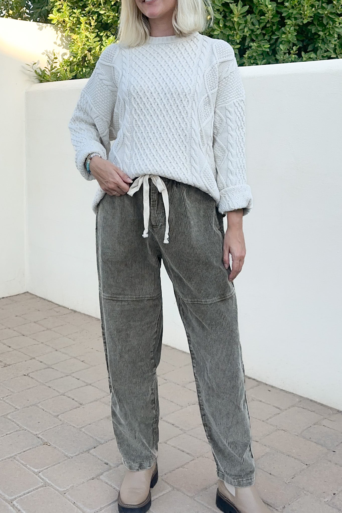 A woman wearing a white cable-knit sweater and gray corduroy drawstring pants, standing outdoors against a white wall with green foliage in the background.