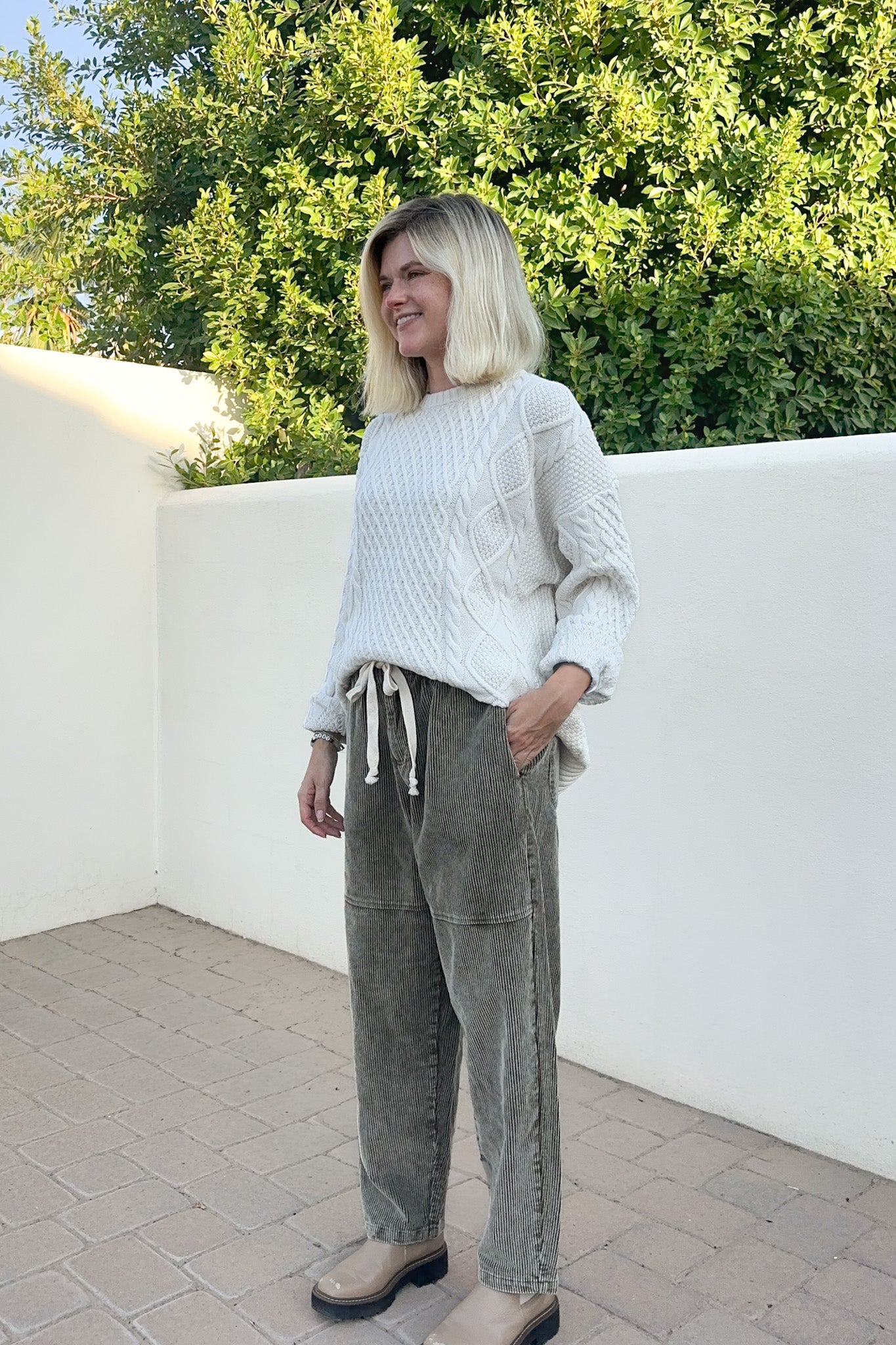 A woman with blonde hair stands outdoors wearing a white cable-knit sweater and olive green corduroy pants with a drawstring waist, paired with beige shoes.