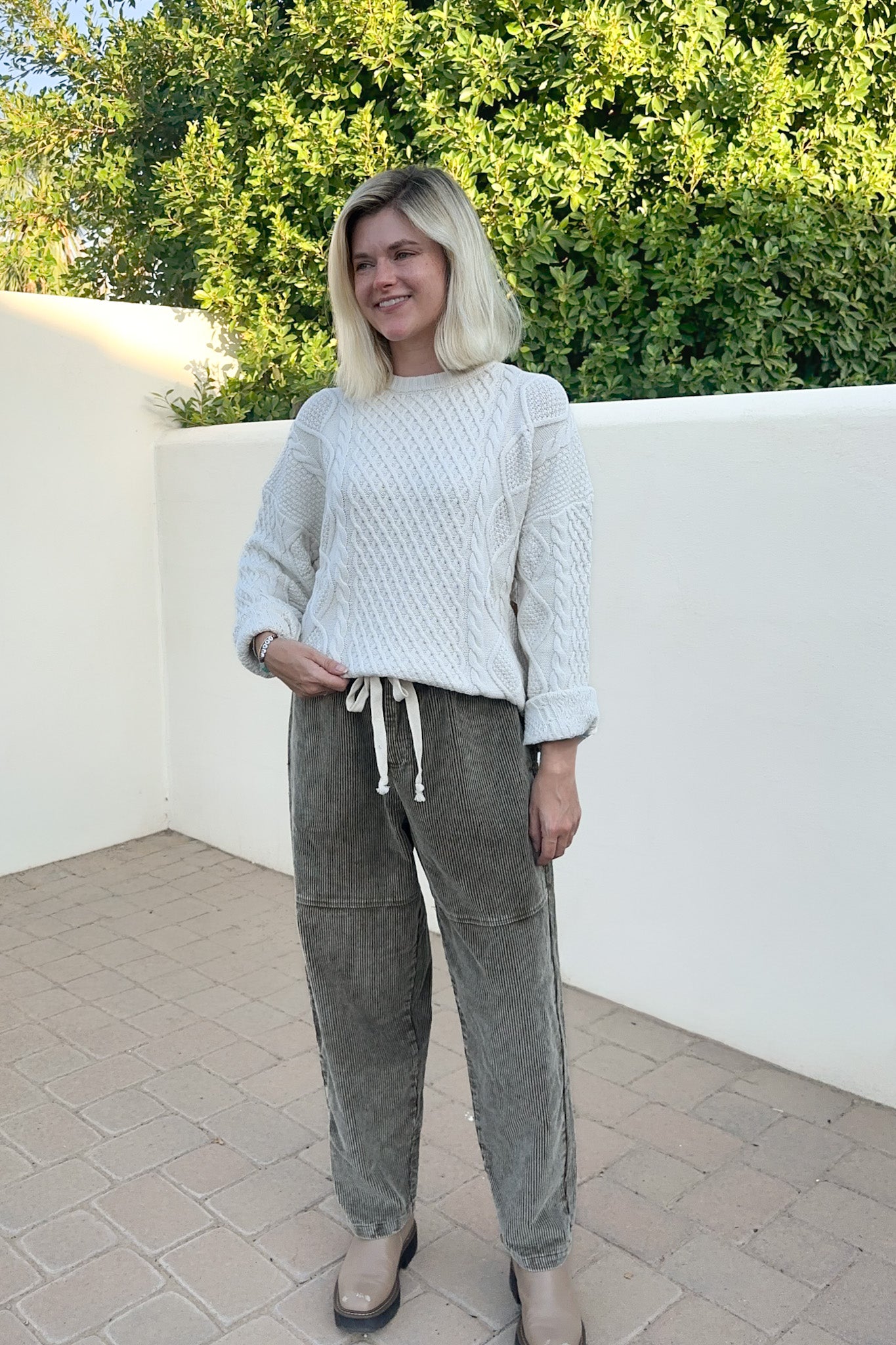 A woman wearing a cream cable-knit sweater and olive green corduroy pants, standing outdoors against a white wall and green foliage.