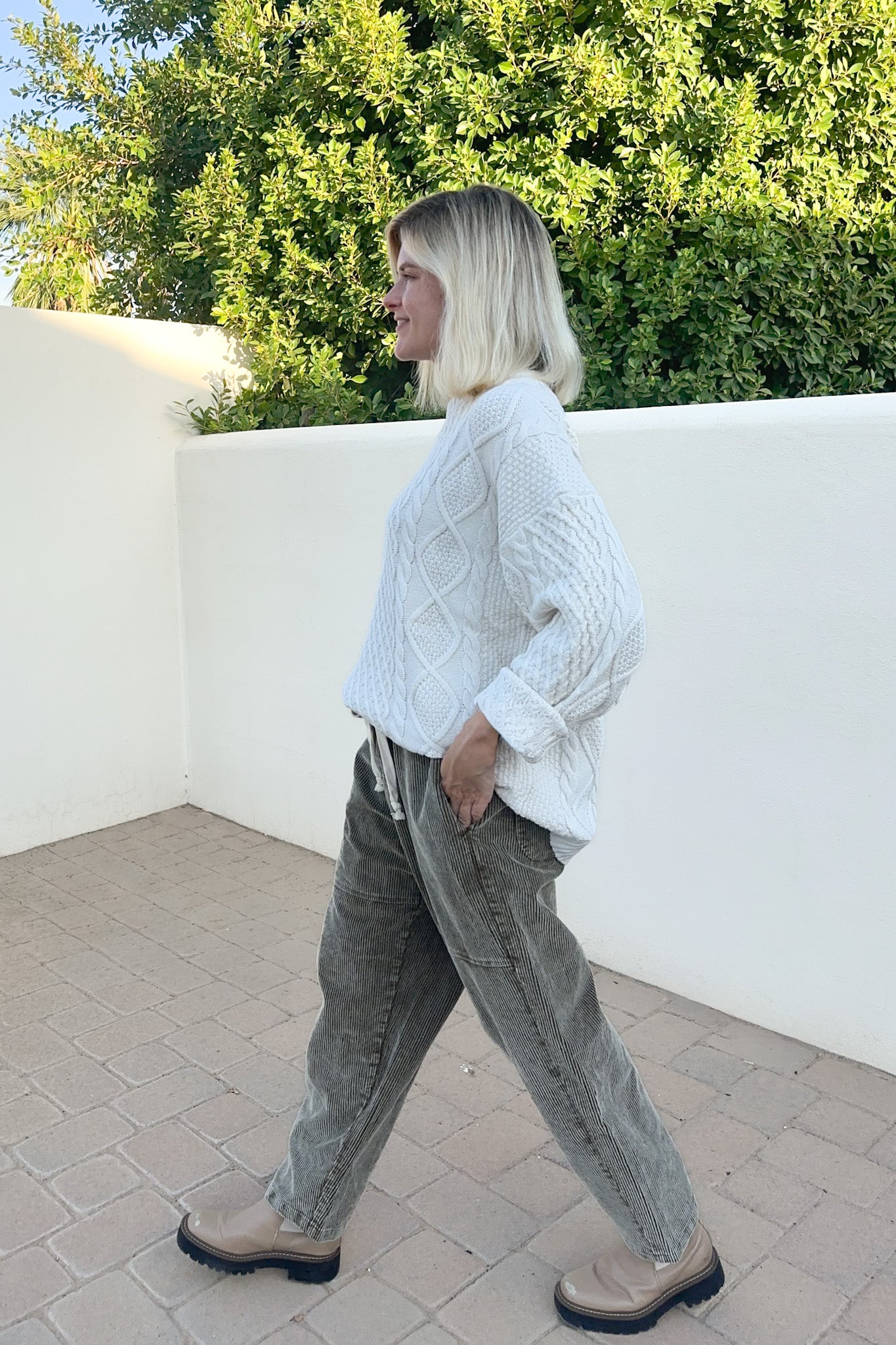 A woman with blonde hair is shown in profile walking outdoors, wearing a white cable-knit sweater, gray corduroy pants, and beige boots. She is standing on a paved patio with a white wall and green foliage in the background.