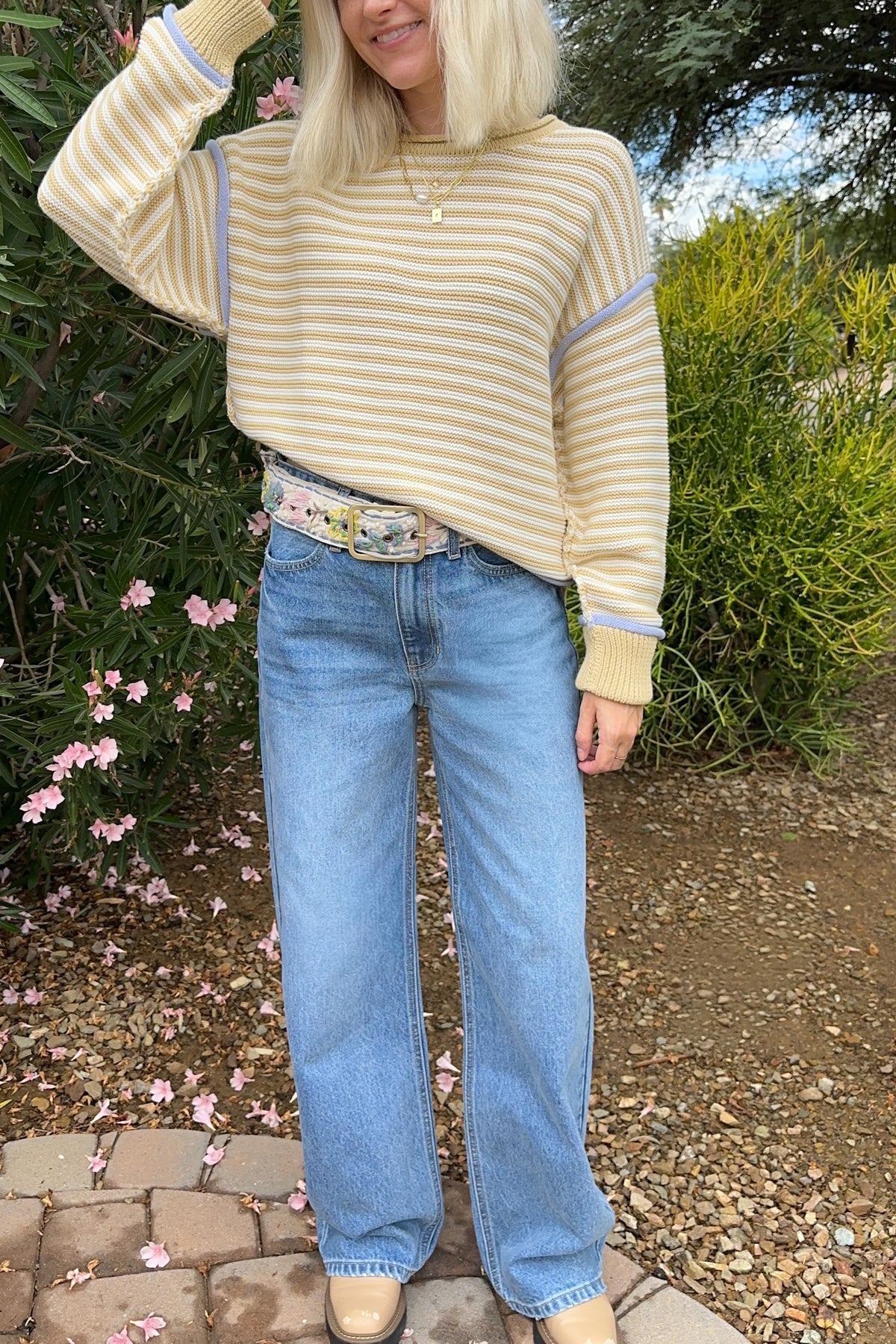 A woman wearing a cream and yellow striped knit sweater with light blue trim, paired with light-wash blue jeans and beige boots, standing outdoors in a garden setting.