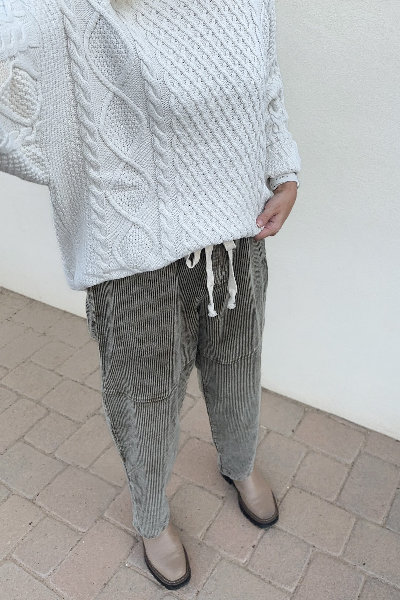 A person wearing a chunky white cable-knit sweater and gray corduroy drawstring pants, standing on a light brick surface against a plain off-white wall.