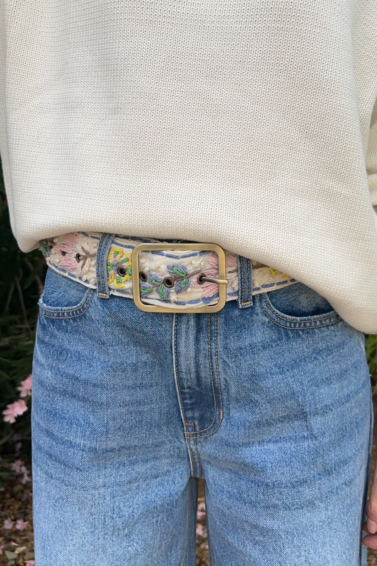 A close-up of a person wearing a light beige knit sweater, blue jeans, and a decorative embroidered belt with a gold-tone buckle. The belt features floral embroidery in pink, yellow, and green on a light base, paired with a large rectangular gold buckle.