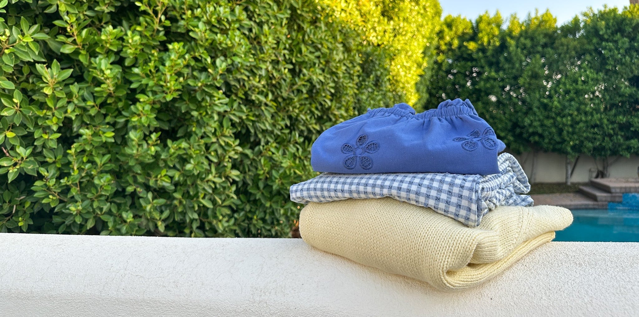 A stack of three folded reusable cloth napkins or washcloths sits on a white outdoor ledge with a lush green hedge and pool area in the background. The top napkin is blue with a subtle floral pattern, the middle is blue and white gingham, and the bottom is a solid pale yellow/beige.