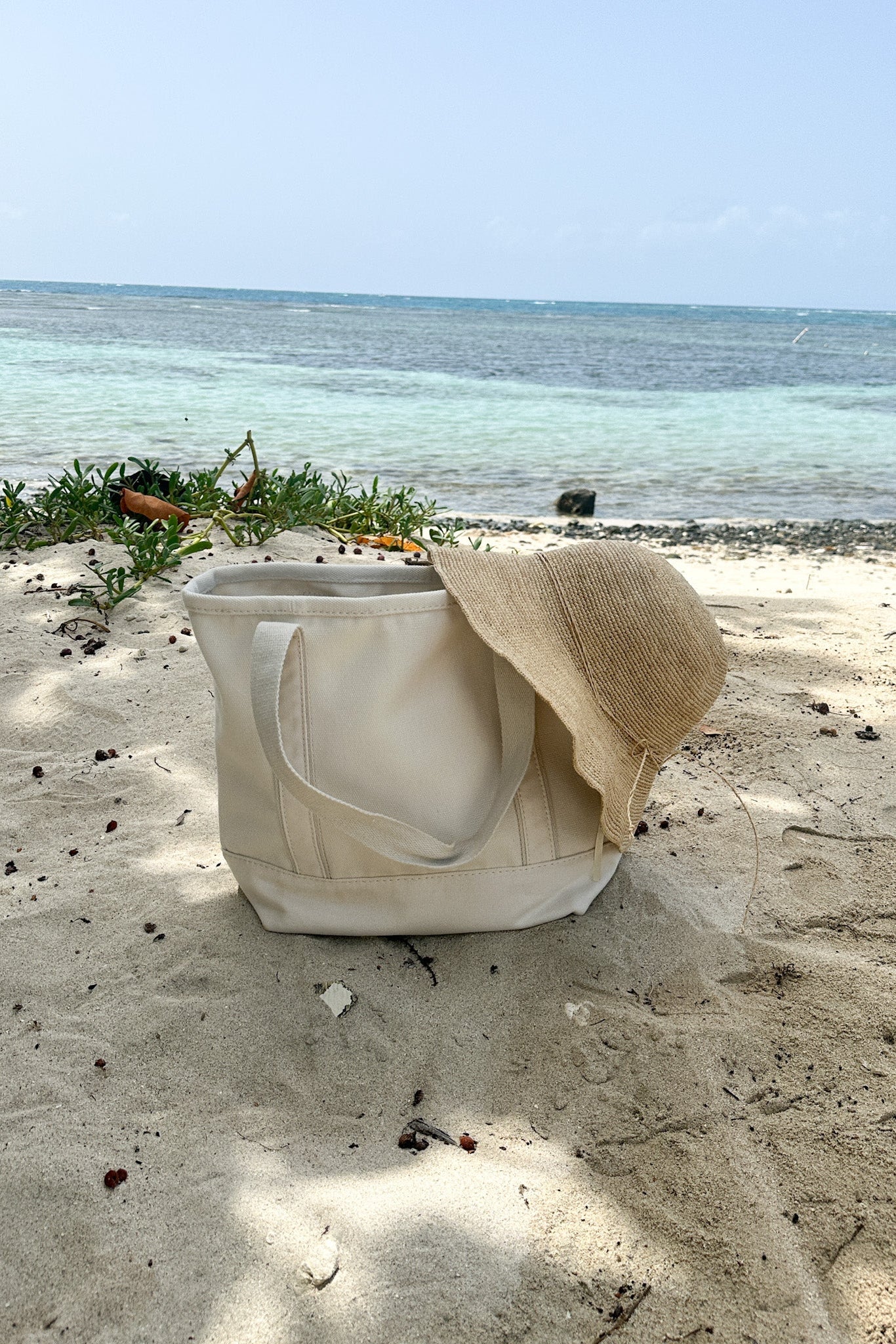 A cream canvas tote bag sits on a sandy beach with the ocean and sky in the background, with a straw hat draped over one of its handles. 