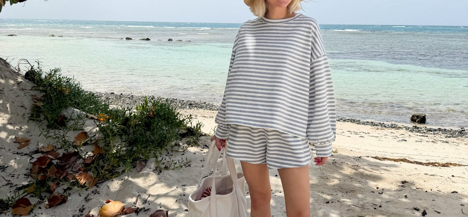 A woman stands on a sandy beach wearing a matching beige and white striped long-sleeve top and shorts set, a wide-brimmed straw hat, and holding a white tote bag.