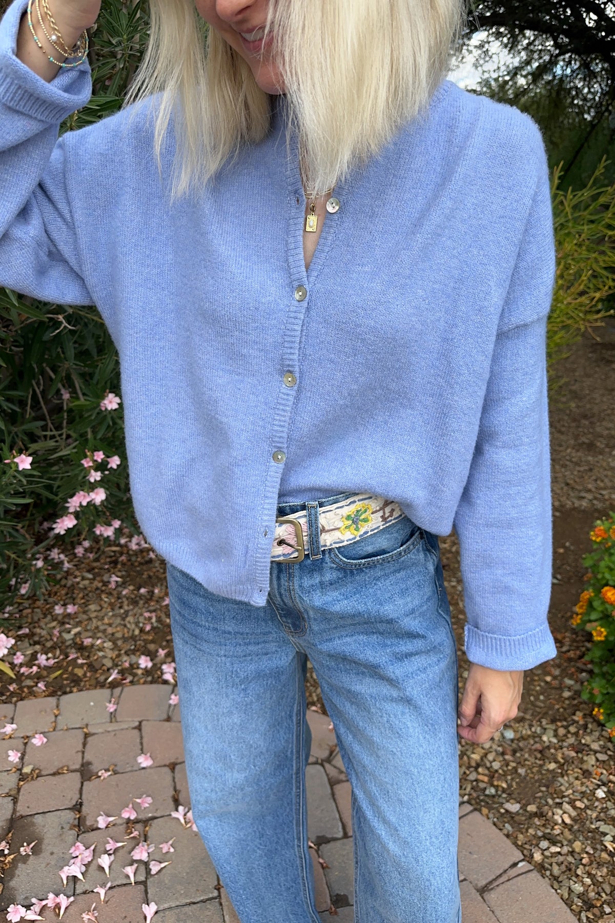 A person outdoors wearing a light blue knit button-front cardigan over a white floral top, paired with light-wash blue jeans and a patterned belt.
