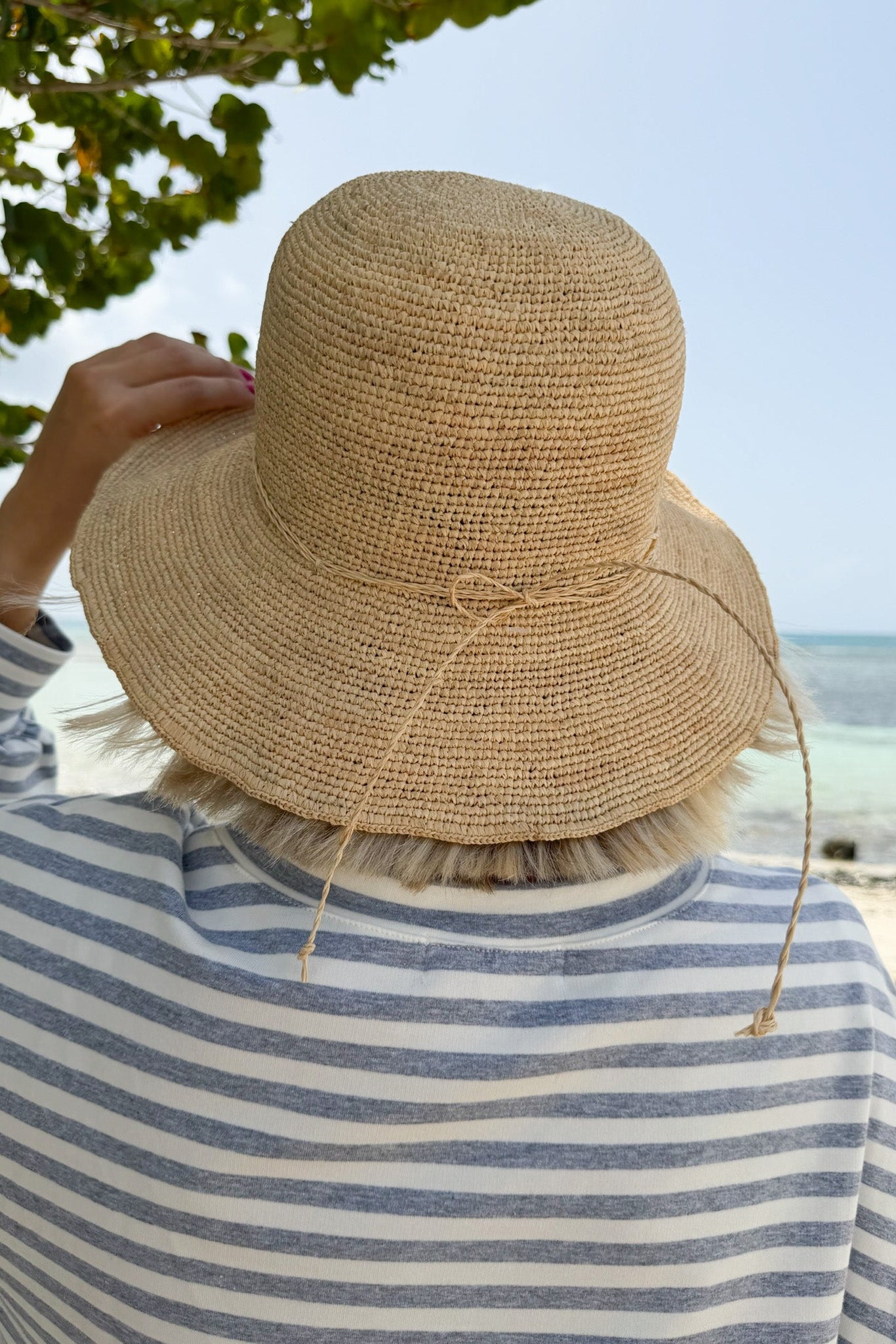 A person viewed from behind wearing a natural straw sun hat with a tie detail and a blue-and-white striped shirt, standing outdoors near a beach with ocean and sky in the background.