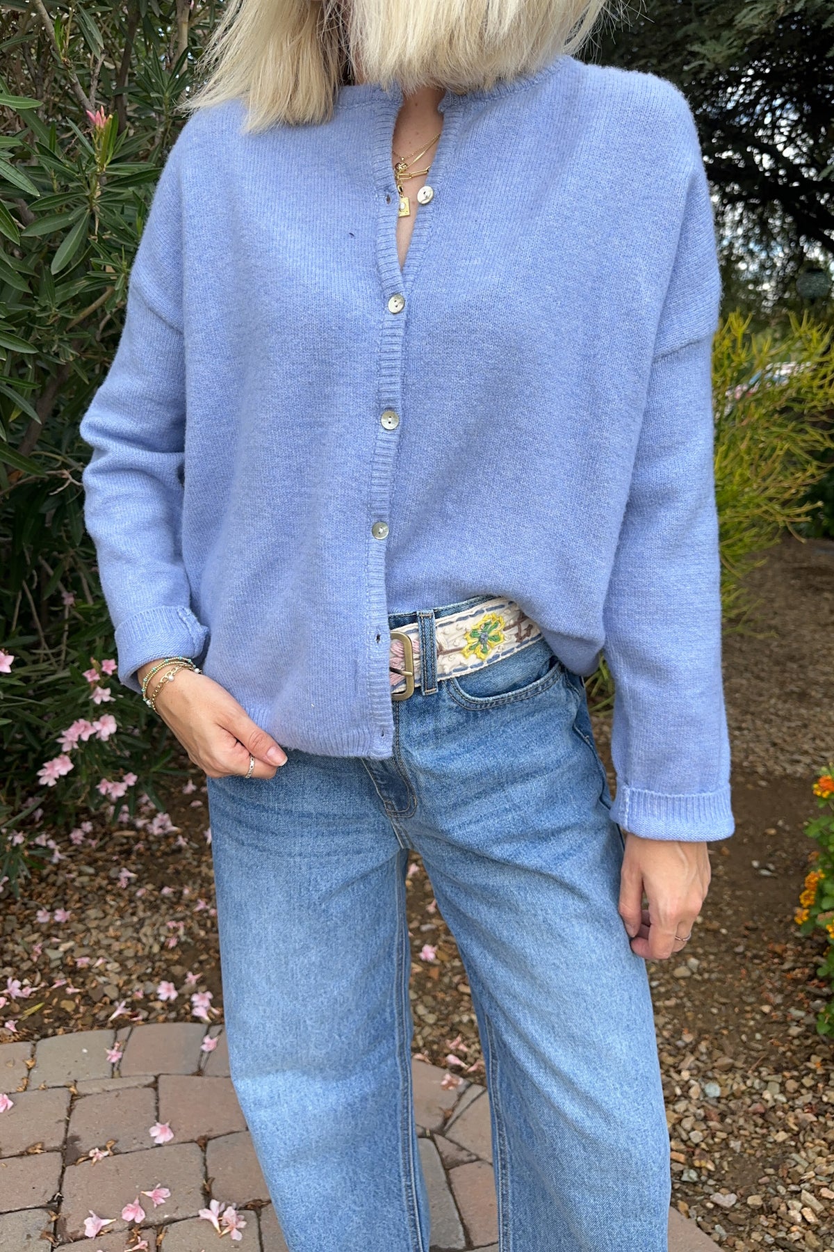 A person wearing a light periwinkle blue knit cardigan with a split neckline and gold-tone buttons, paired with light-wash blue jeans in an outdoor garden setting.