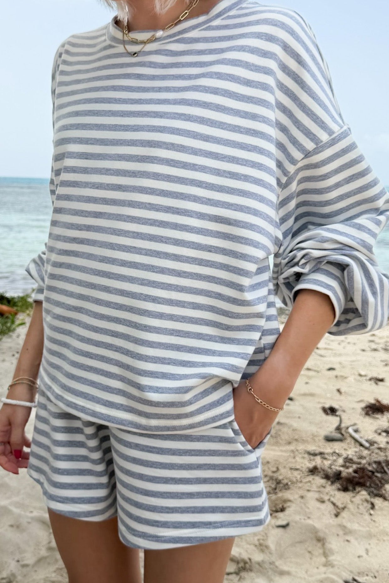 A person standing on a sandy beach wearing an oversized long-sleeve striped top and matching shorts in light blue and white.