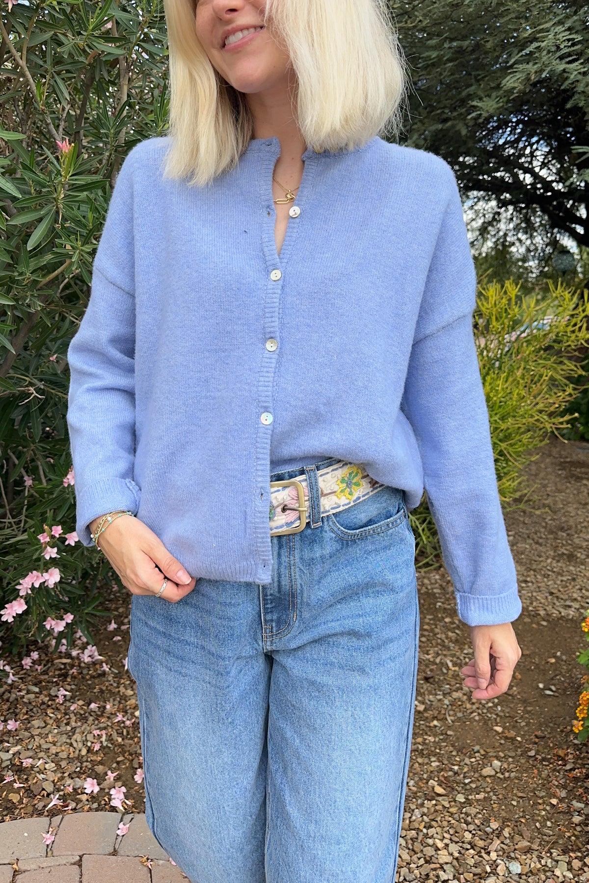 A woman wearing a light blue knit cardigan with a buttoned stand collar, paired with light-wash blue jeans and a floral belt, standing outdoors in front of green garden foliage.