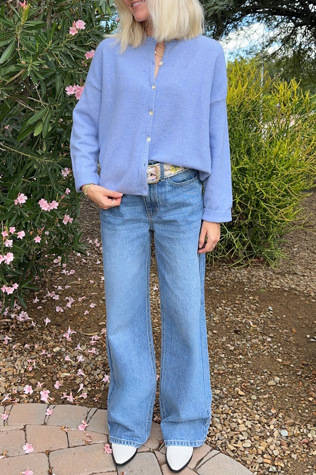 A woman wearing a light periwinkle blue cropped button-up knit cardigan and wide-leg light blue jeans, standing outdoors in a garden setting.