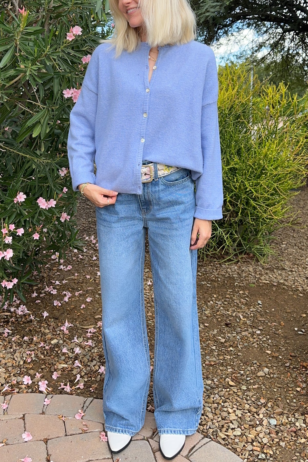 A woman wearing a light periwinkle blue cropped button-up knit cardigan and wide-leg light blue jeans, standing outdoors in a garden setting.