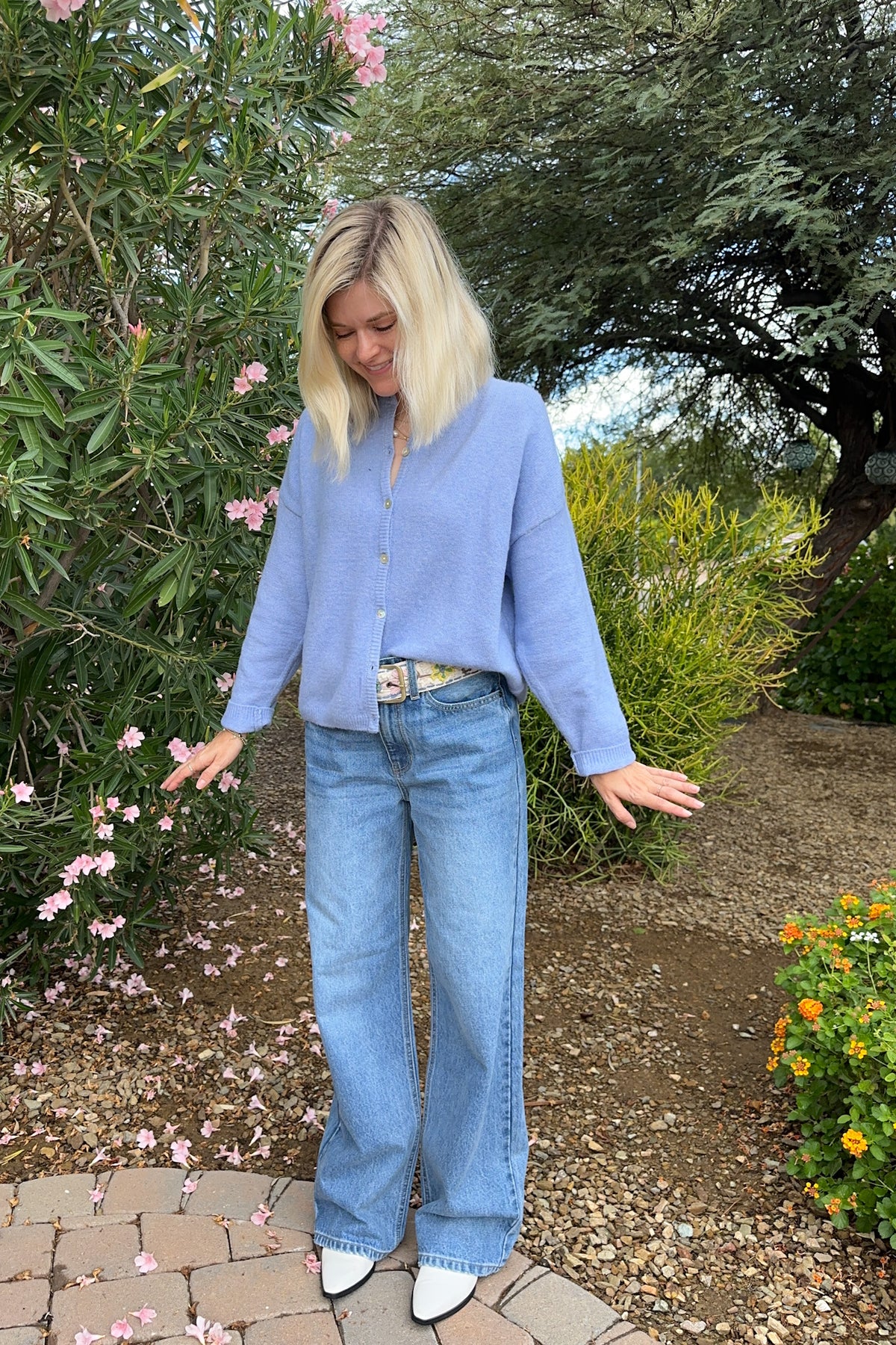 A woman stands outdoors in a garden wearing a light periwinkle blue knit cardigan, wide-leg light blue jeans, and white shoes.