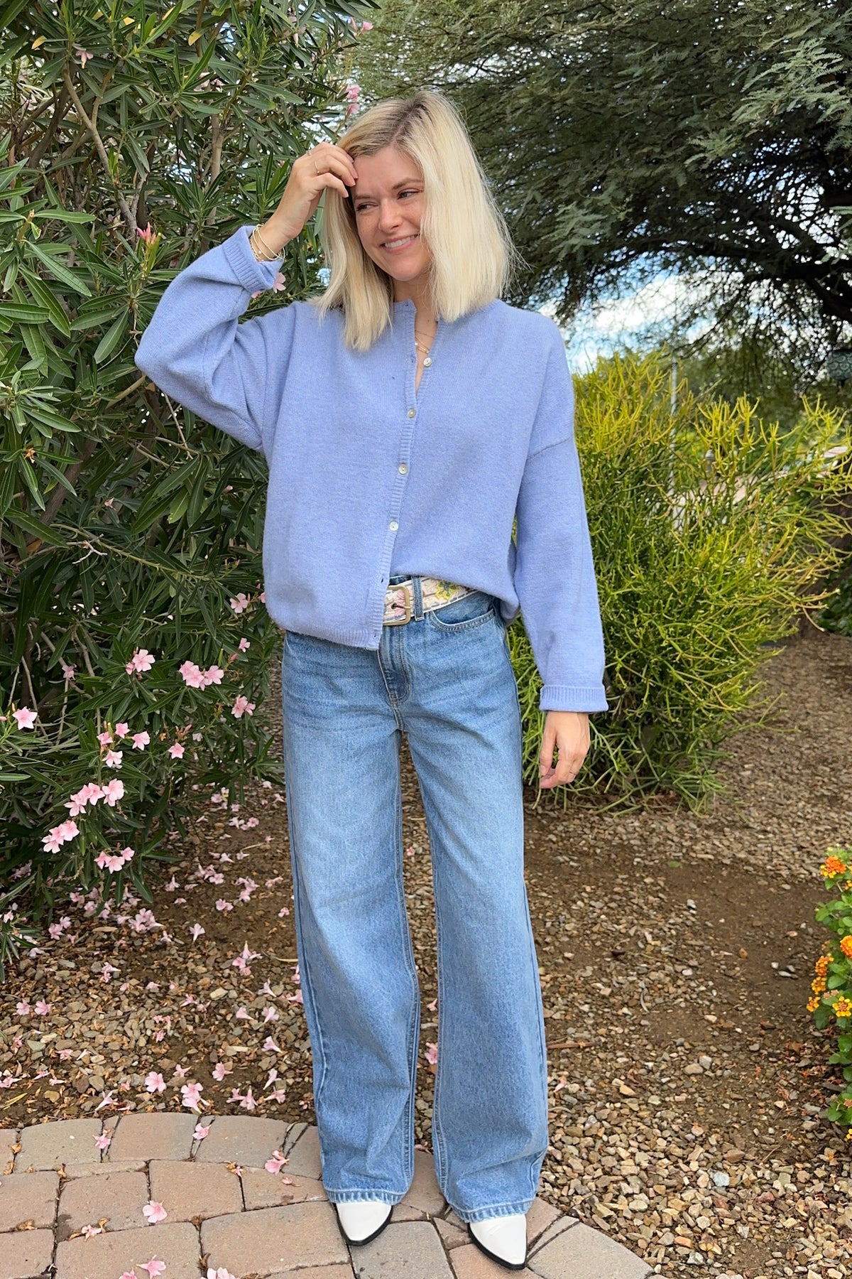 A woman with blonde hair stands outdoors in a garden setting, wearing a light blue long-sleeve button-up shirt, light blue wide-leg jeans, and white pointed shoes.