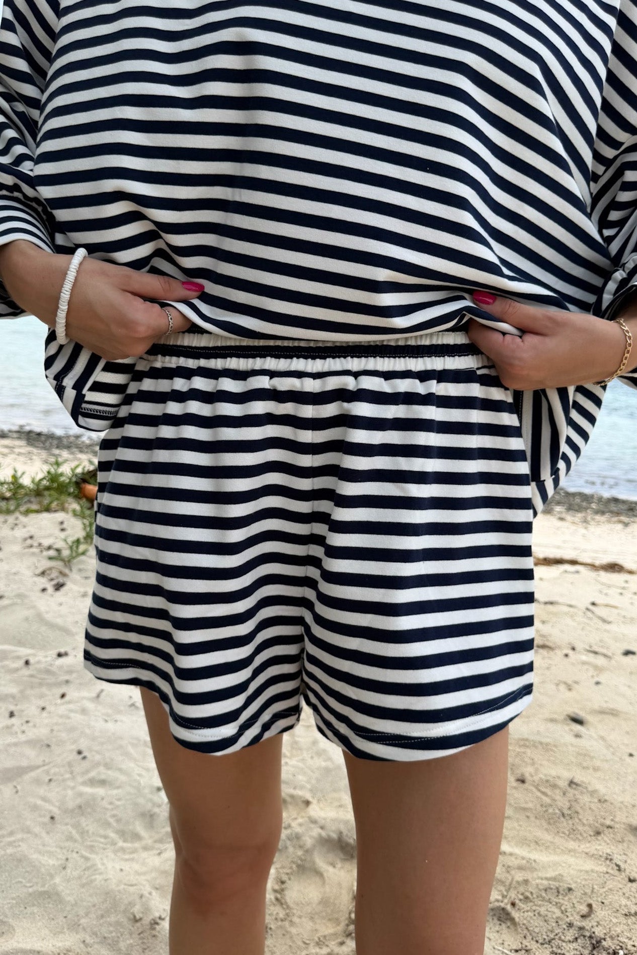 A person standing on a sandy beach wearing a matching navy and white horizontally striped lounge set consisting of a long-sleeve top and shorts.