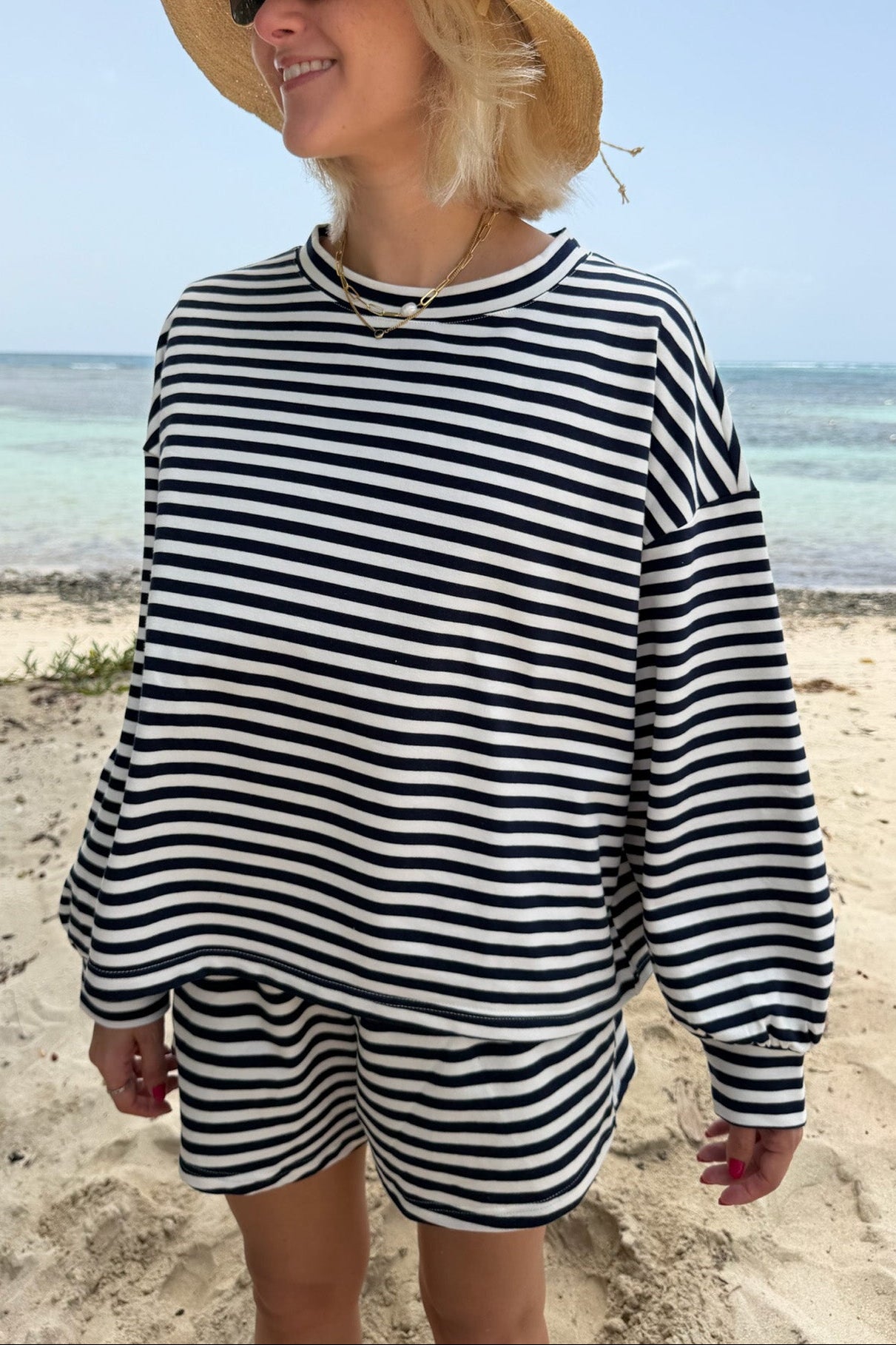 A woman standing on a sandy beach wearing a navy and white horizontally striped long-sleeve top and matching shorts, with a straw sun hat and sunglasses.