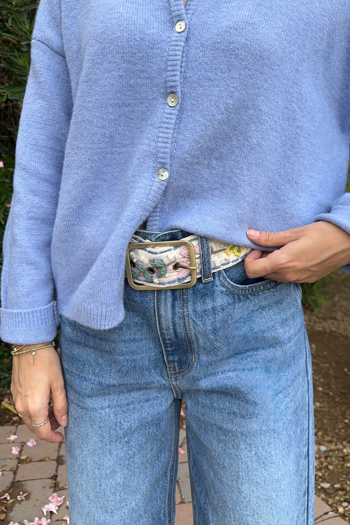 A person wearing a light blue knit cardigan and light blue jeans, with the cardigan tucked into a belt featuring a colorful floral print and gold-tone buckle.