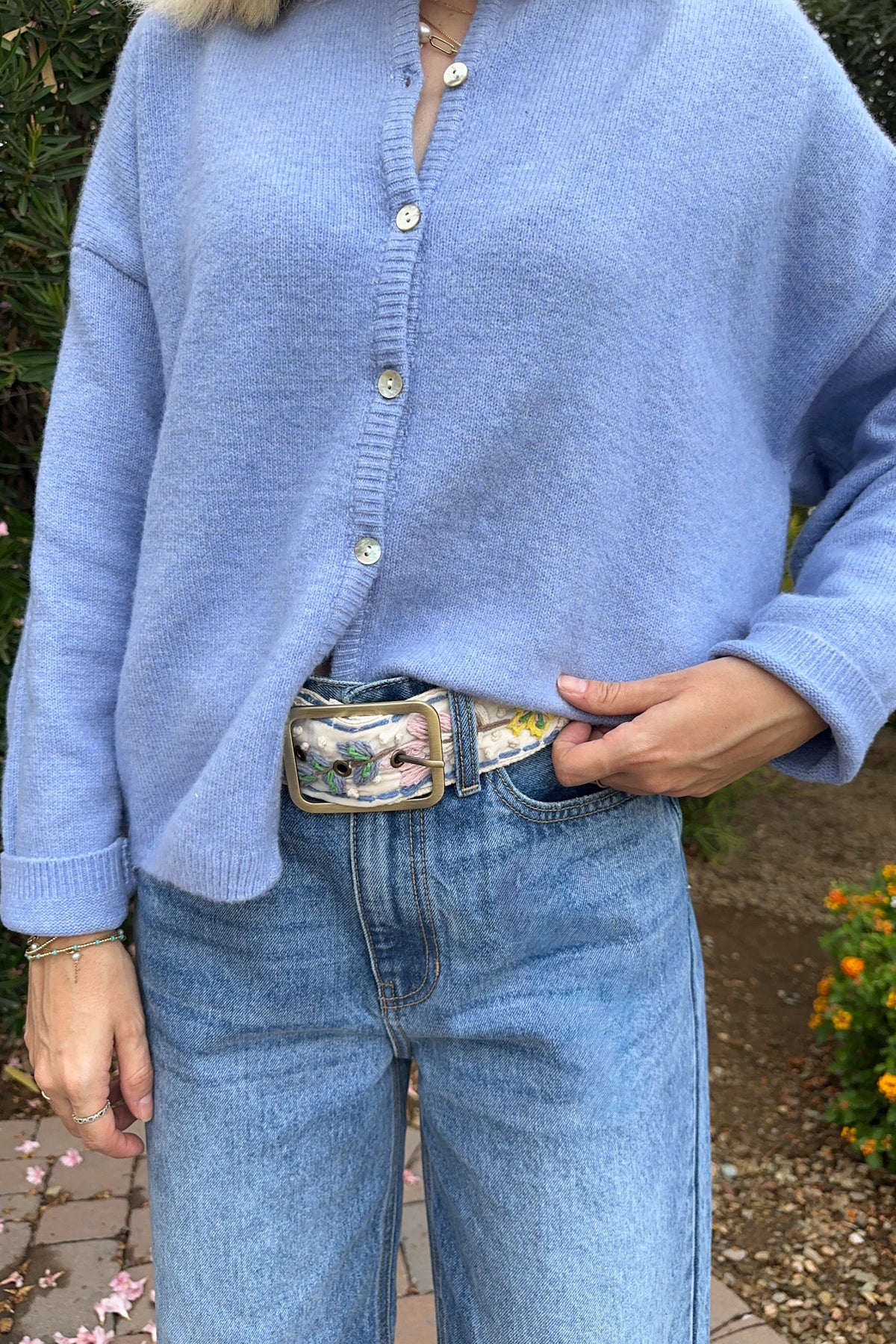A person wearing a light blue knit button-front cardigan and light blue jeans, standing outdoors with one hand in a pocket. The cardigan has a soft fuzzy texture and small light buttons, paired with a patterned belt.