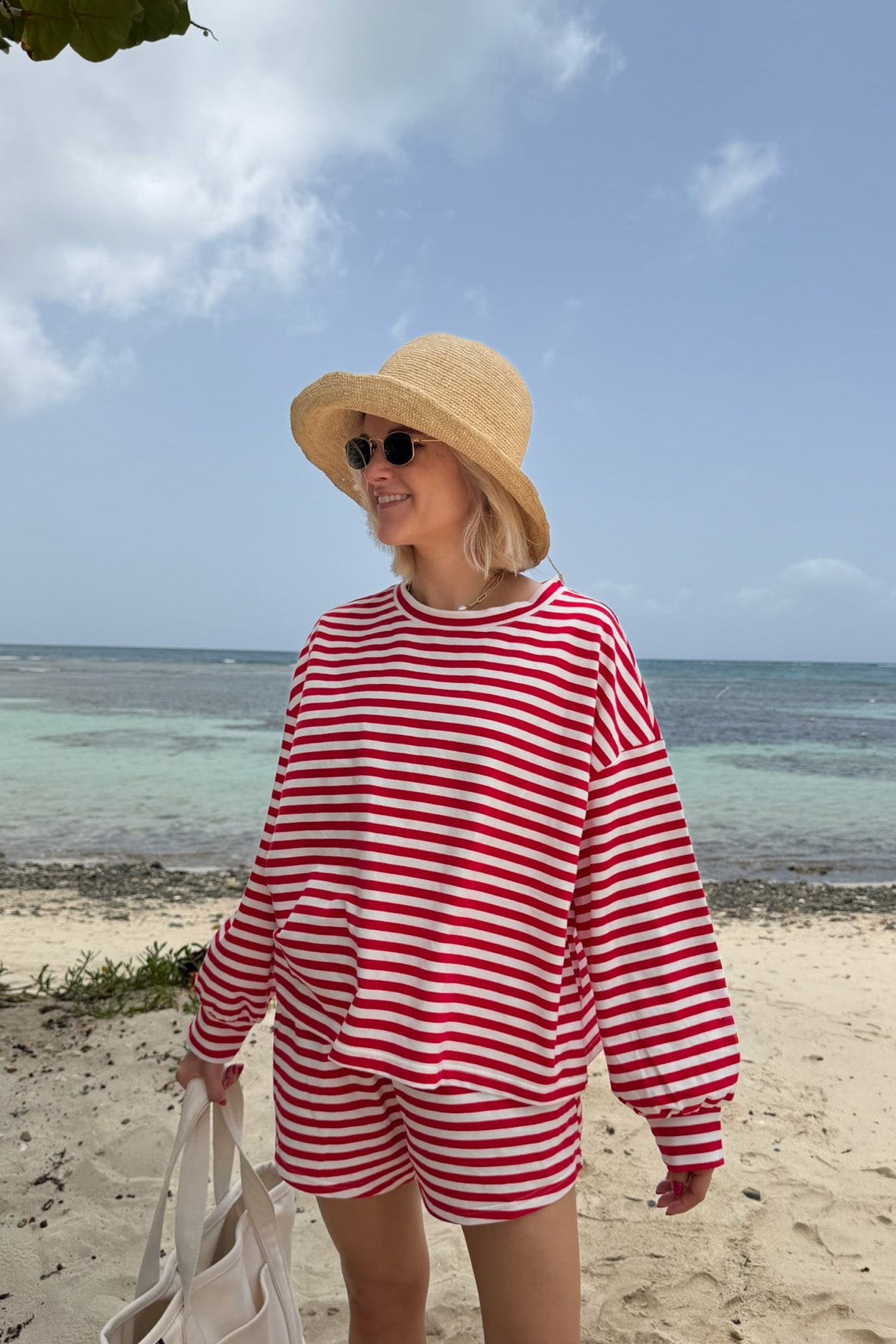 A woman standing on a sandy beach wearing a red and white striped long-sleeve top and matching shorts, a straw sun hat, sunglasses, and holding a cream tote bag.
