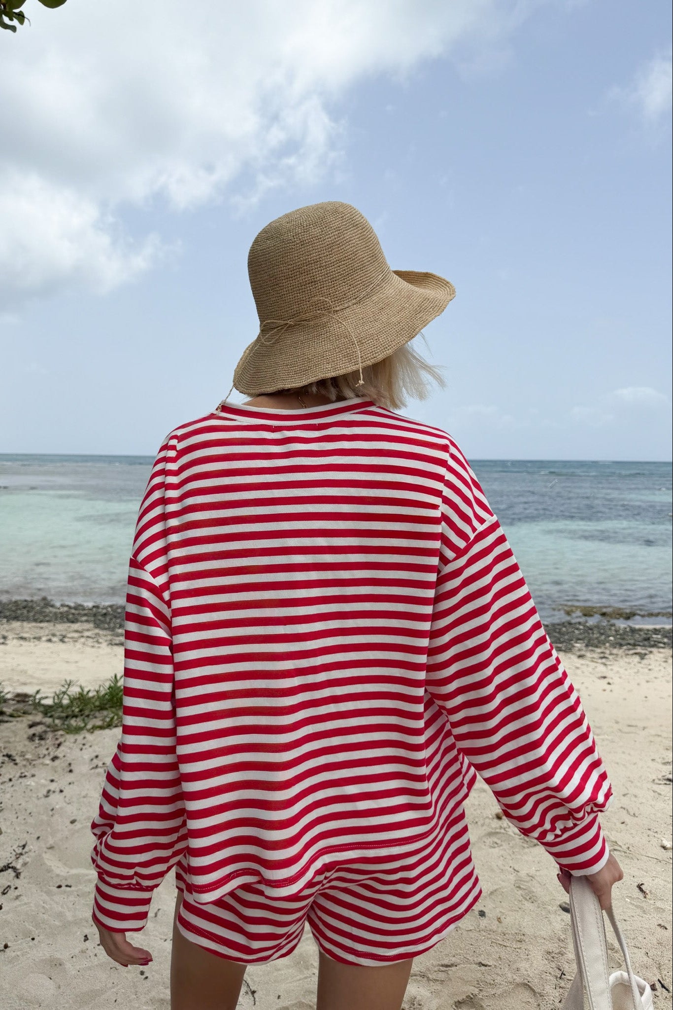 A woman viewed from behind on a sandy beach, wearing a red and white horizontally striped long-sleeve top and matching shorts, with a beige straw sun hat.