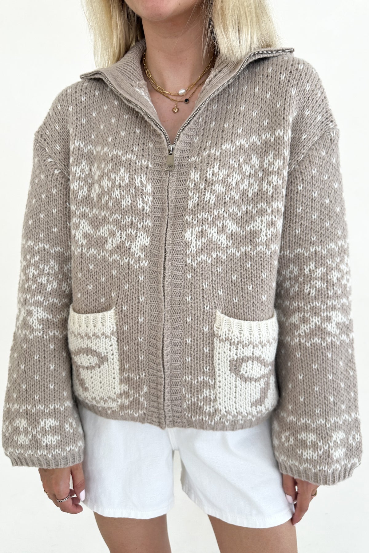 A woman wearing a beige and white patterned knit zip-up cardigan sweater with pockets, paired with white shorts, against a plain white background.