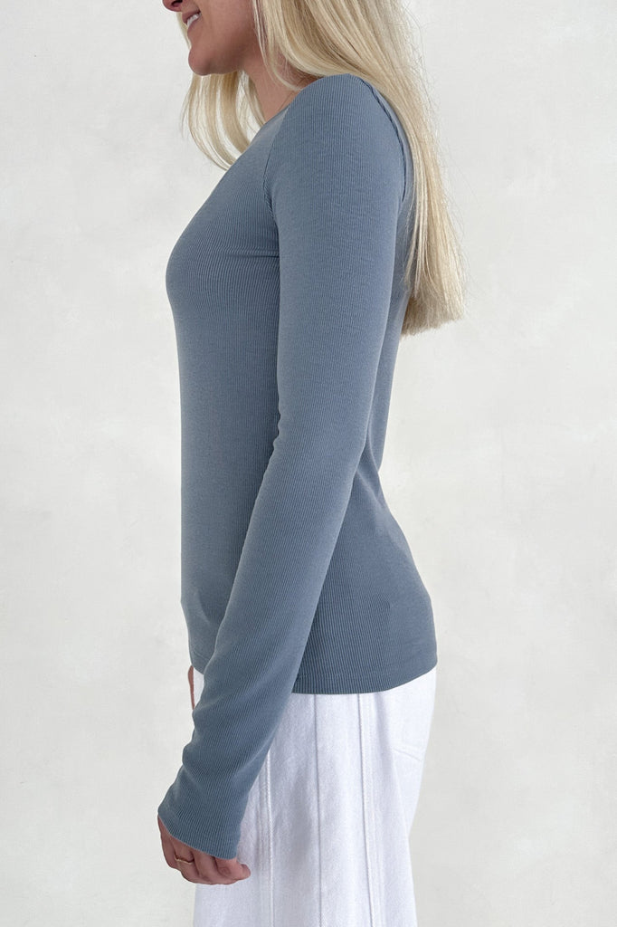 Woman wearing a blue long-sleeve top and white pants on a light gray background