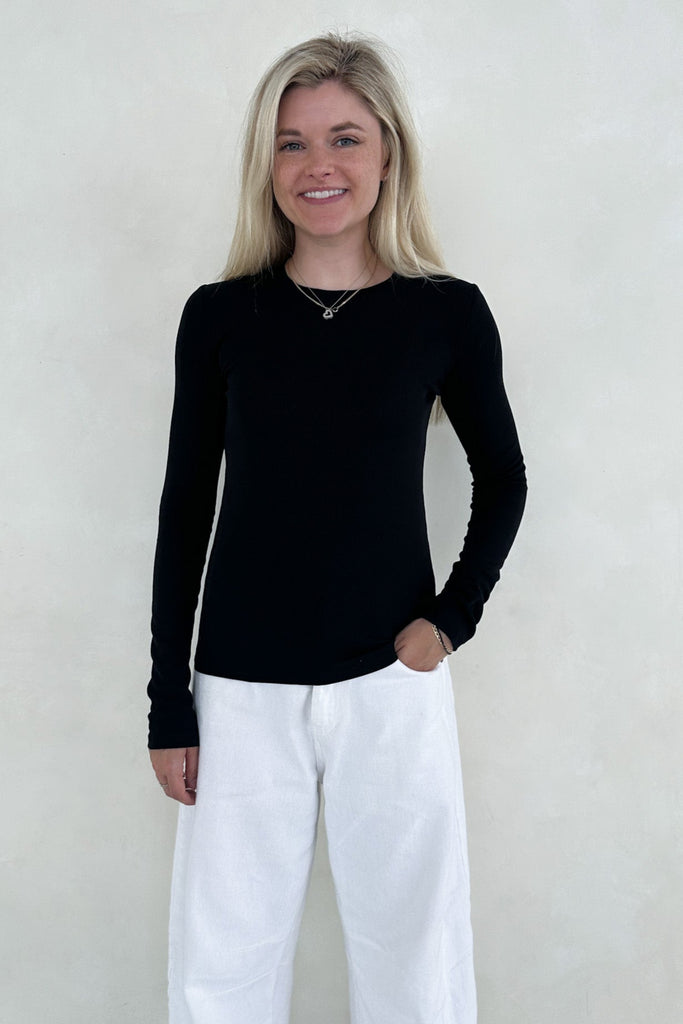 Woman wearing a black long-sleeve top and white pants against a plain background