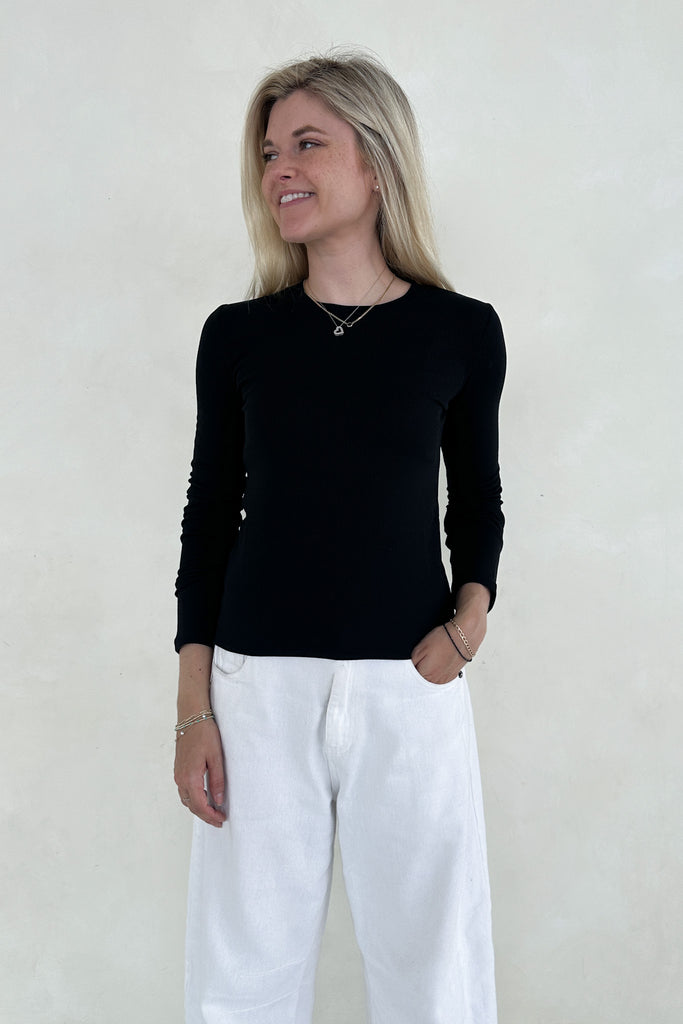 Woman wearing a black long-sleeve top and white pants against a plain background