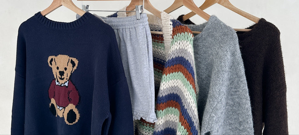 Collection of sweaters and pants on hangers against a white background