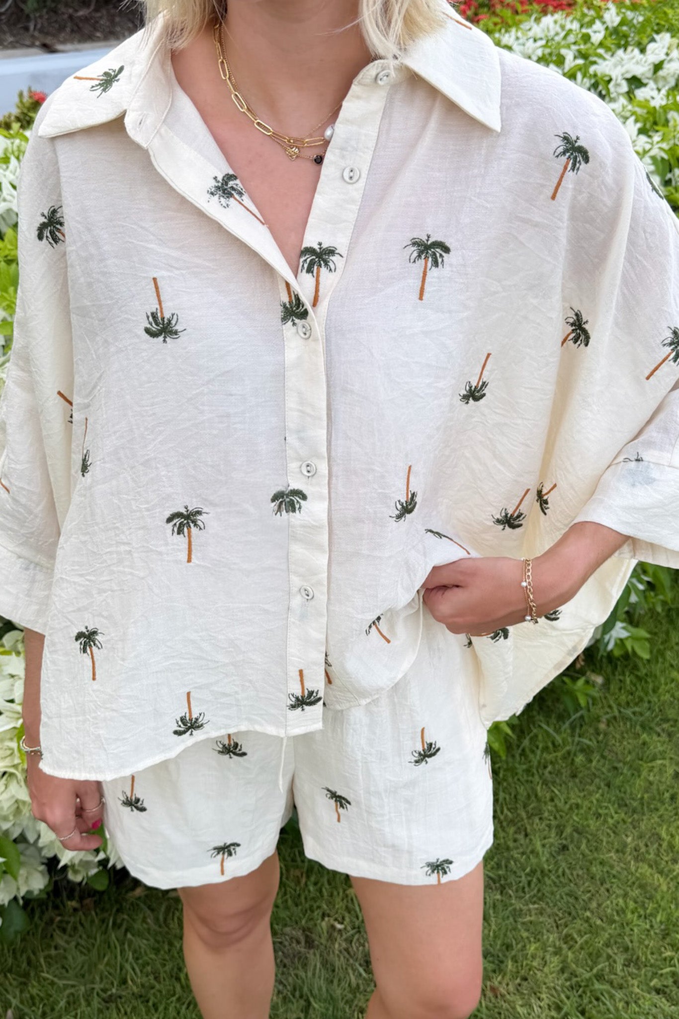 A person wearing a cream-colored linen button-up shirt and matching shorts set, both printed with small embroidered palm trees, photographed outdoors on a grassy lawn.