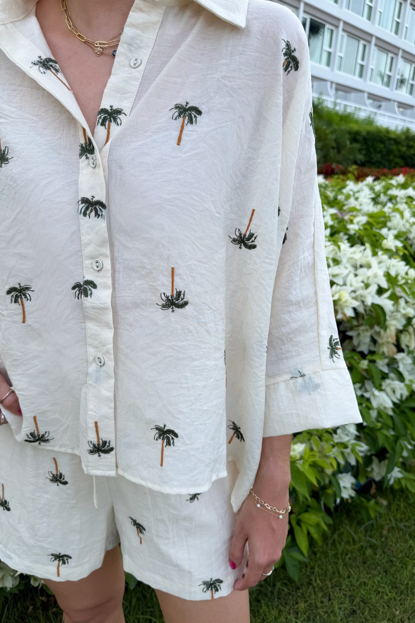 A person wearing a white short-sleeve button-up shirt and matching shorts set, both printed with small embroidered palm tree motifs, photographed outdoors in front of greenery and a building.