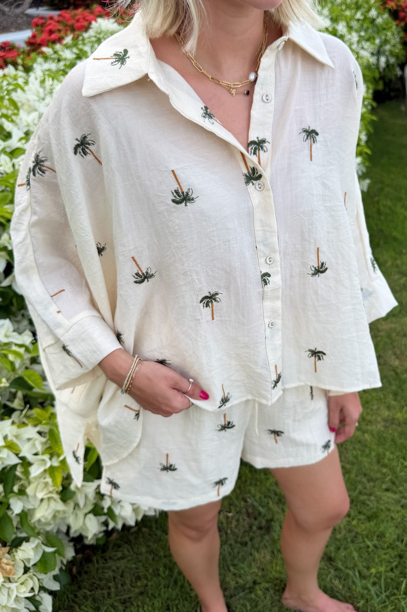 A woman wearing a cream-colored linen shirt and matching shorts set, both decorated with small embroidered palm tree motifs, standing outdoors on a grassy lawn with flowers in the background.