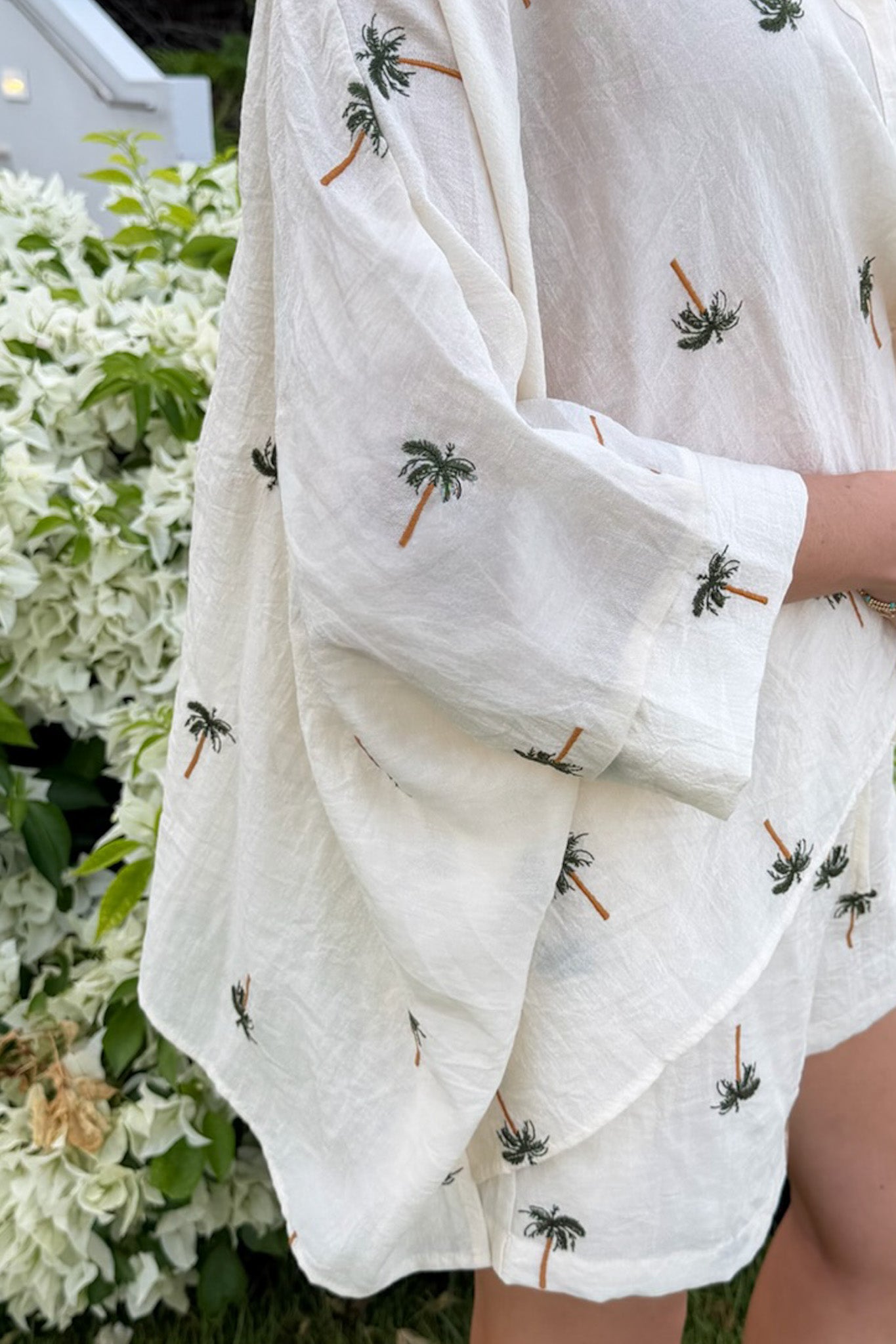 A close-up of a person wearing a loose white cotton cover-up or kimono-style top with small embroidered palm tree motifs, photographed outdoors in front of green foliage.