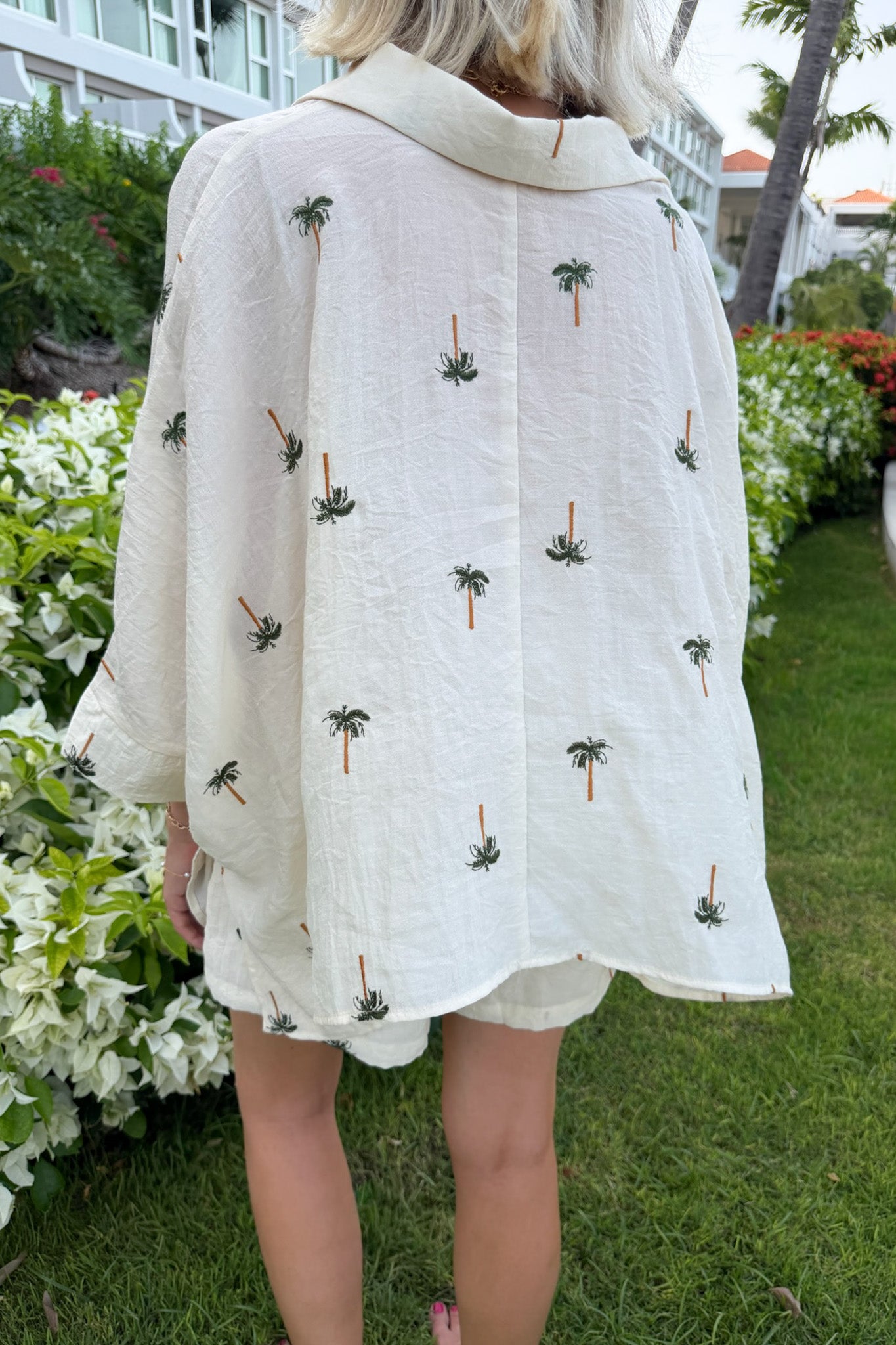 A woman viewed from behind wearing a loose, short-sleeve cream-colored linen shirt with small embroidered palm tree motifs, standing outdoors on a grassy lawn with plants and a building in the background.