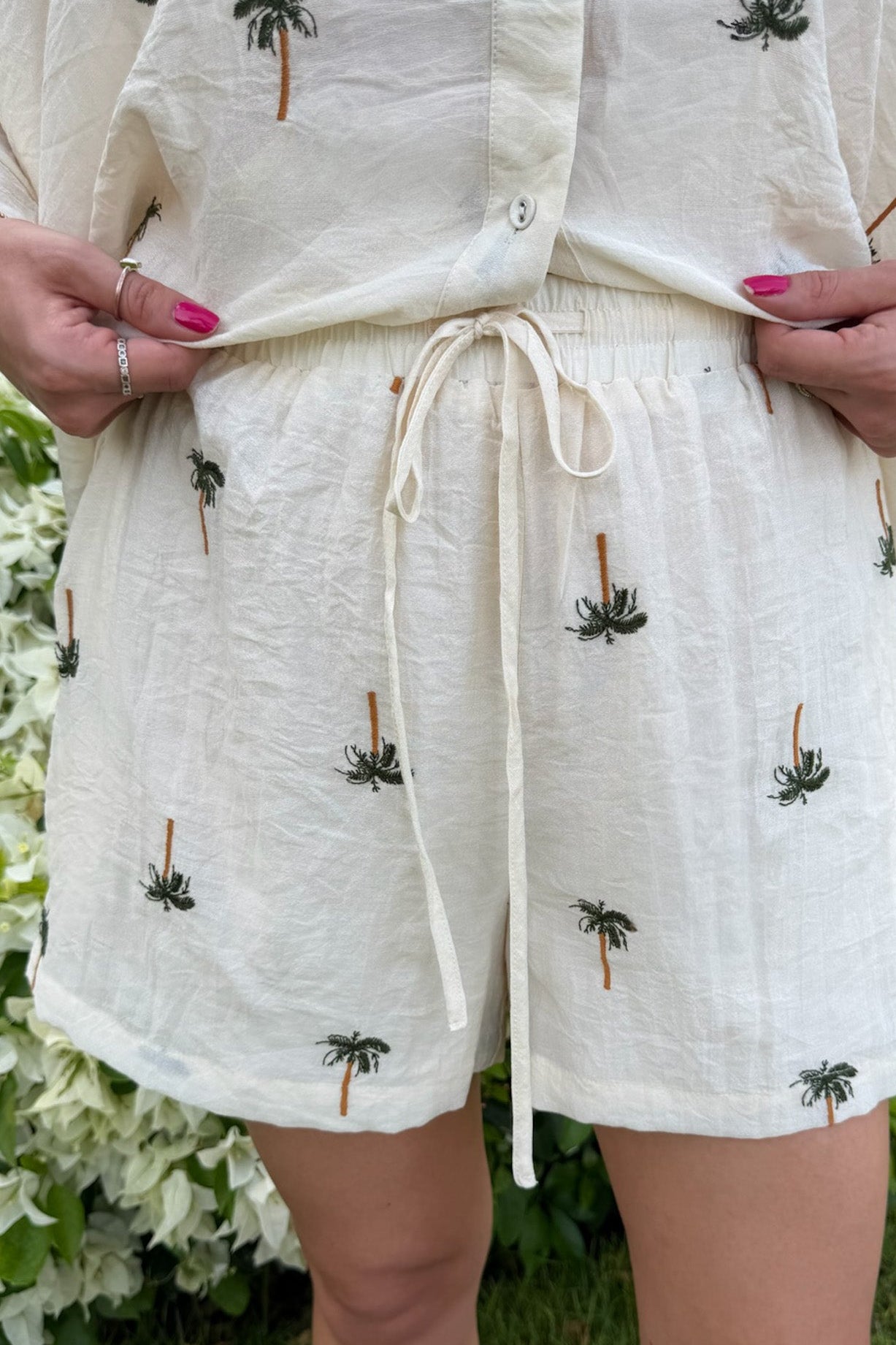 A person wearing a cream-colored linen romper with embroidered palm tree motifs, standing outdoors in front of green foliage and white flowers.