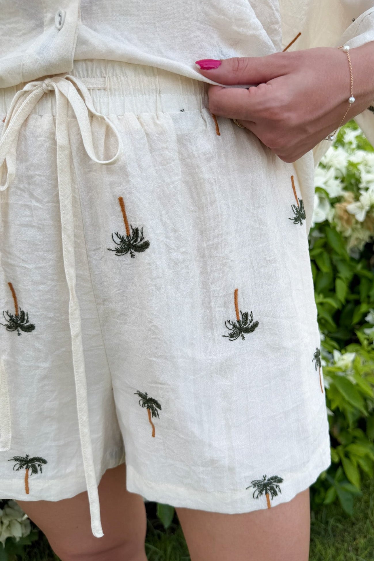 Close-up of a person wearing white linen shorts with embroidered palm tree motifs, standing outdoors in front of green foliage.