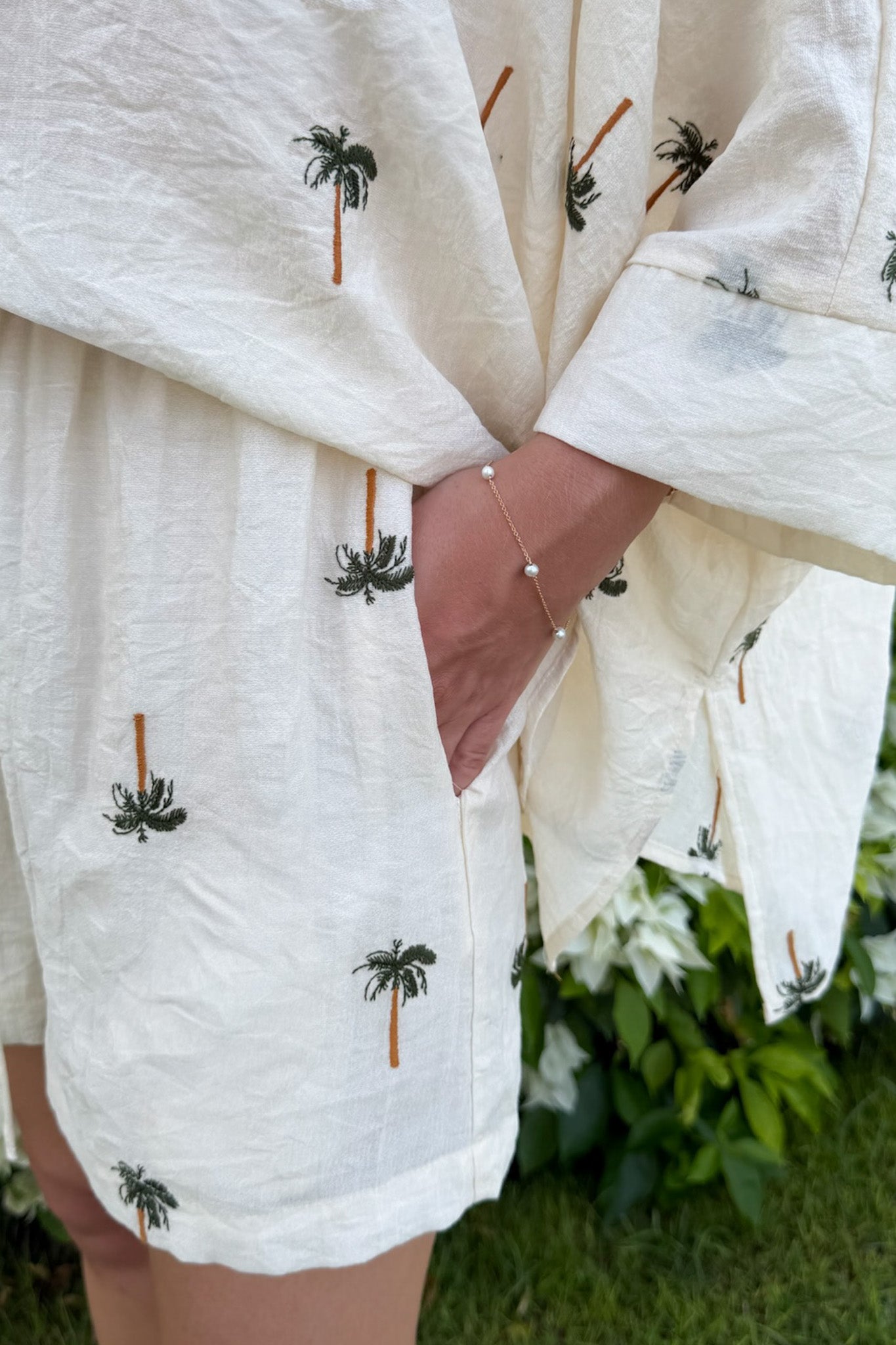 A close-up of a person wearing a loose white linen kimono-style cover-up with small embroidered palm tree motifs, with a hand in a pocket and green foliage in the background.