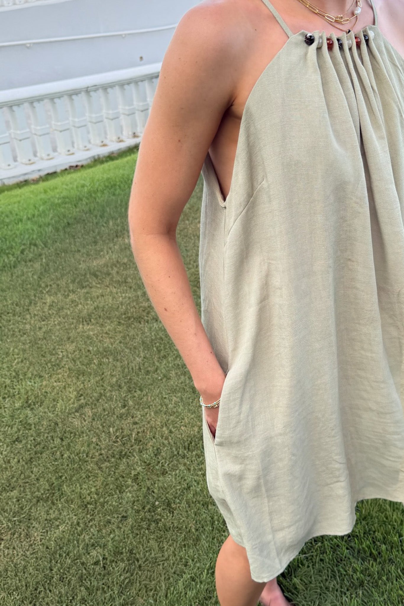 A woman wearing a light beige linen halter dress with side pockets, standing on a green grass lawn.