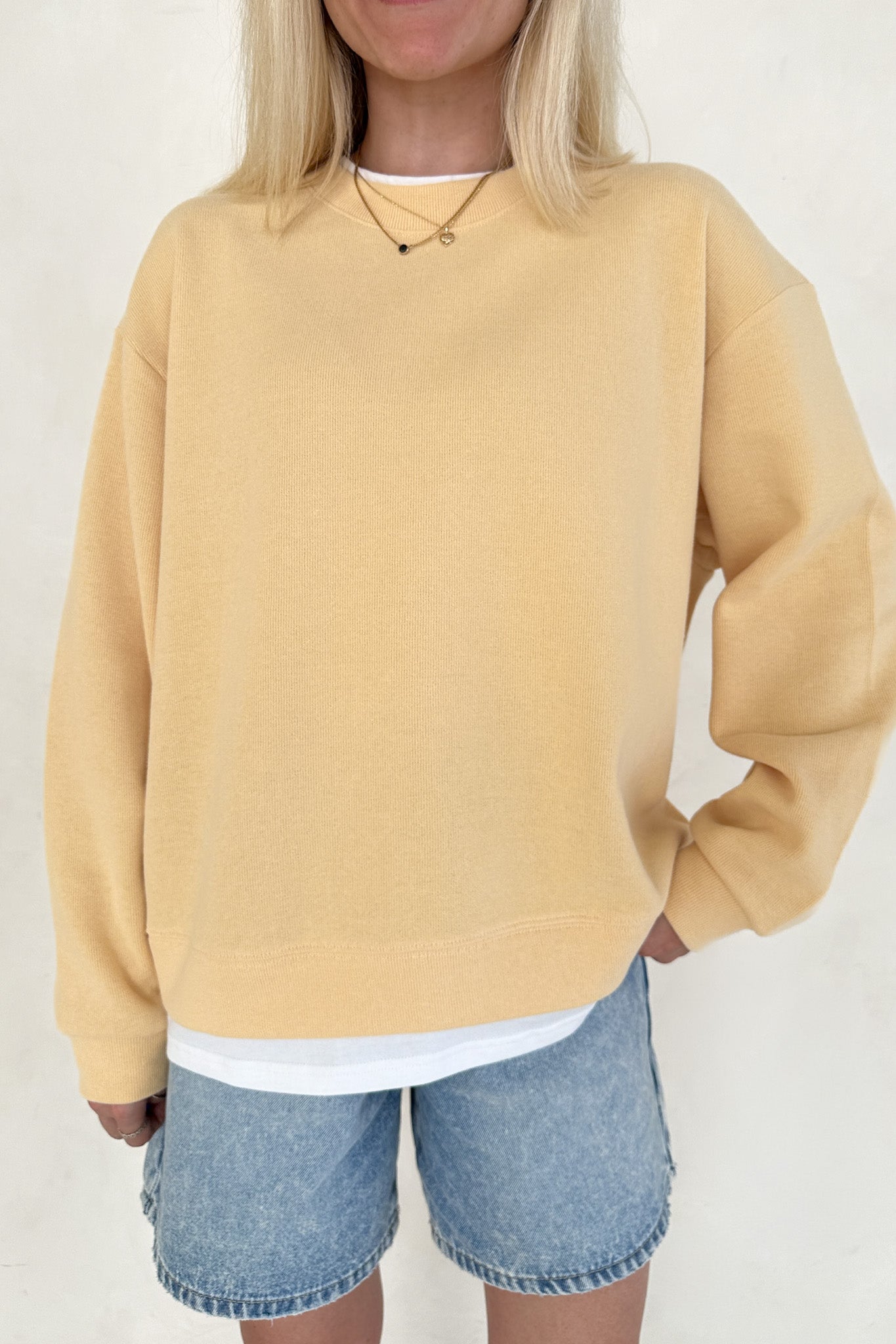 Person wearing a mustard yellow sweatshirt and denim shorts on a plain background