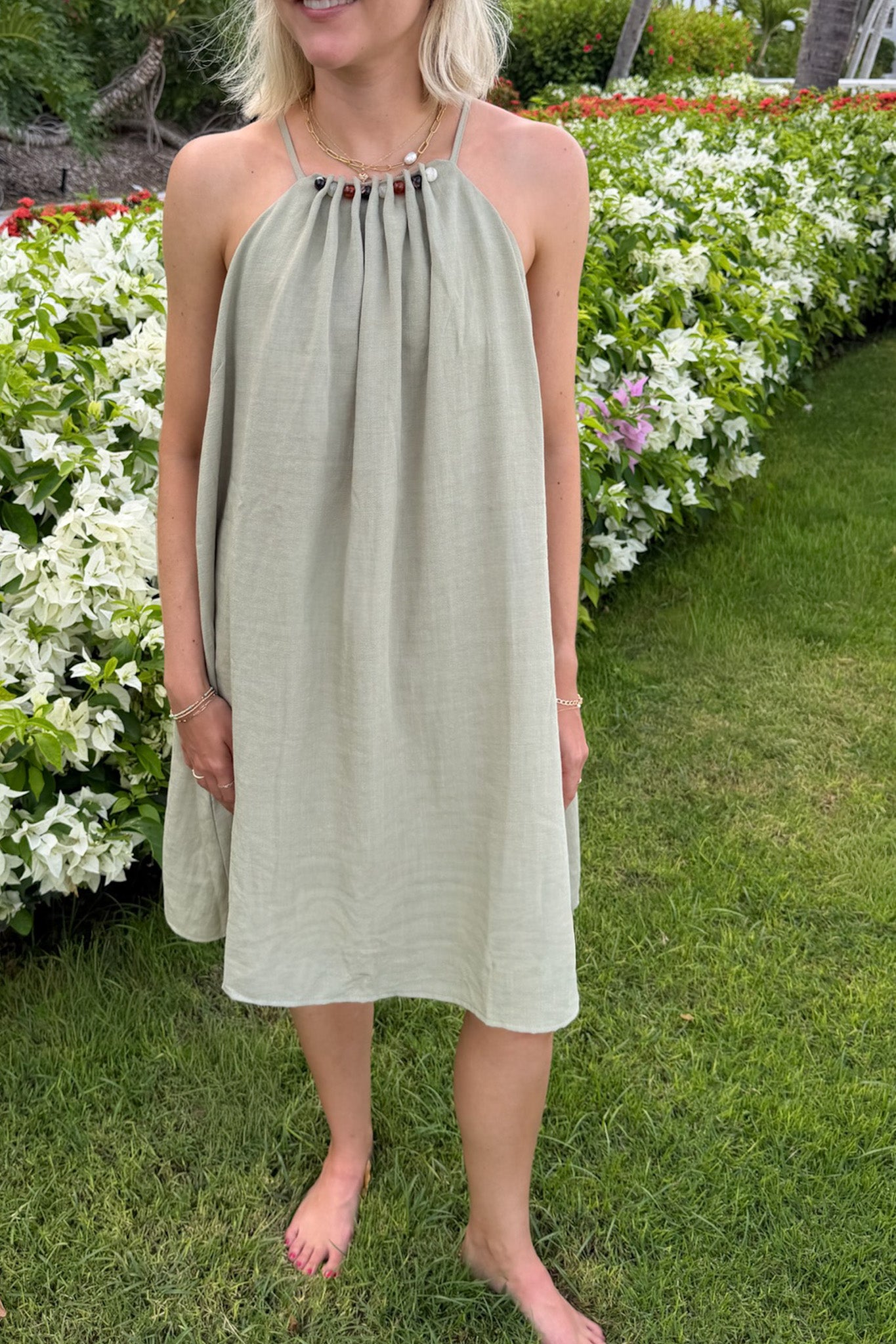 A woman wearing a light sage green linen halter dress with a gathered neckline and small decorative beads, standing barefoot on a grassy lawn with white flowers in the background.