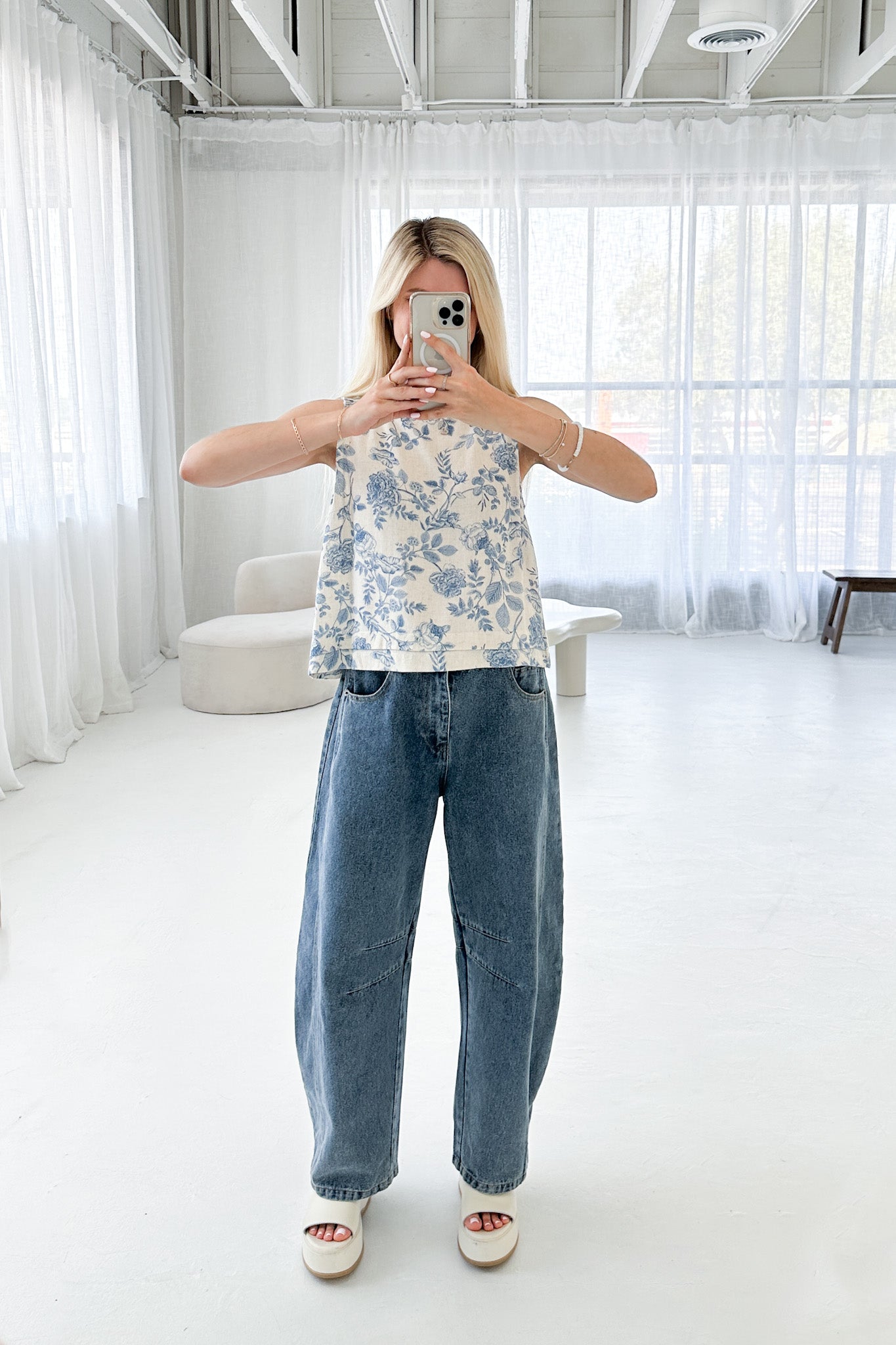 Person taking a mirror selfie wearing a floral top and blue jeans in a minimalistic room.
