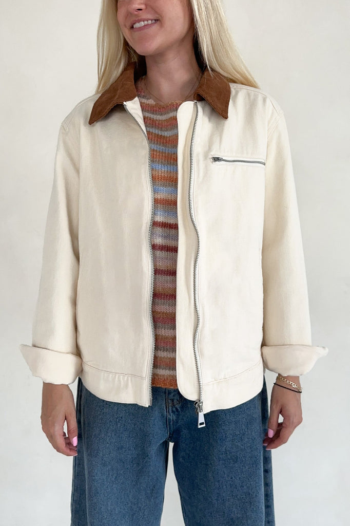 Person wearing a cream jacket with a brown collar, striped sweater, and blue jeans on a white background