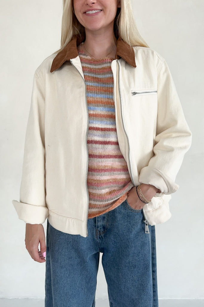 Person wearing a cream jacket, striped sweater, and blue jeans against a white background