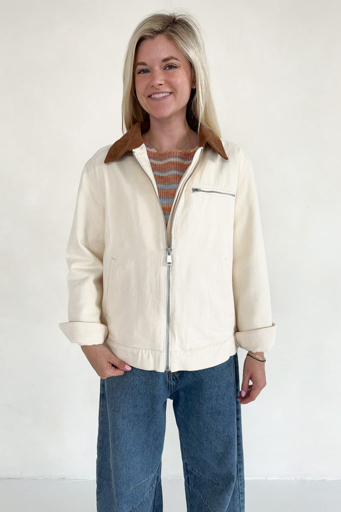 Woman wearing a cream jacket with a brown collar and blue jeans on a white background